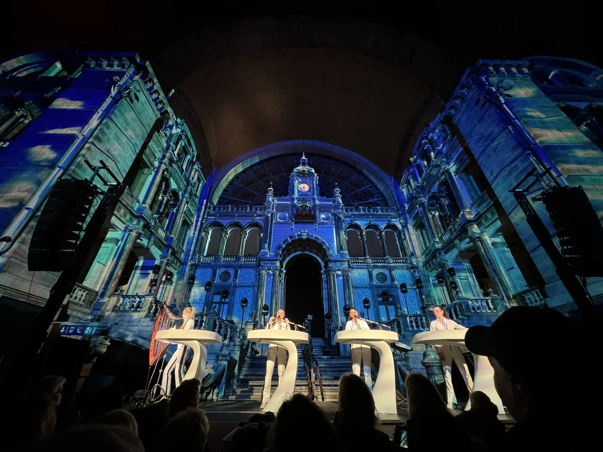 A stage with four performers in front of a large, ornate digital backdrop of a historic building, illuminated in blue light, during a live music or theatrical performance.