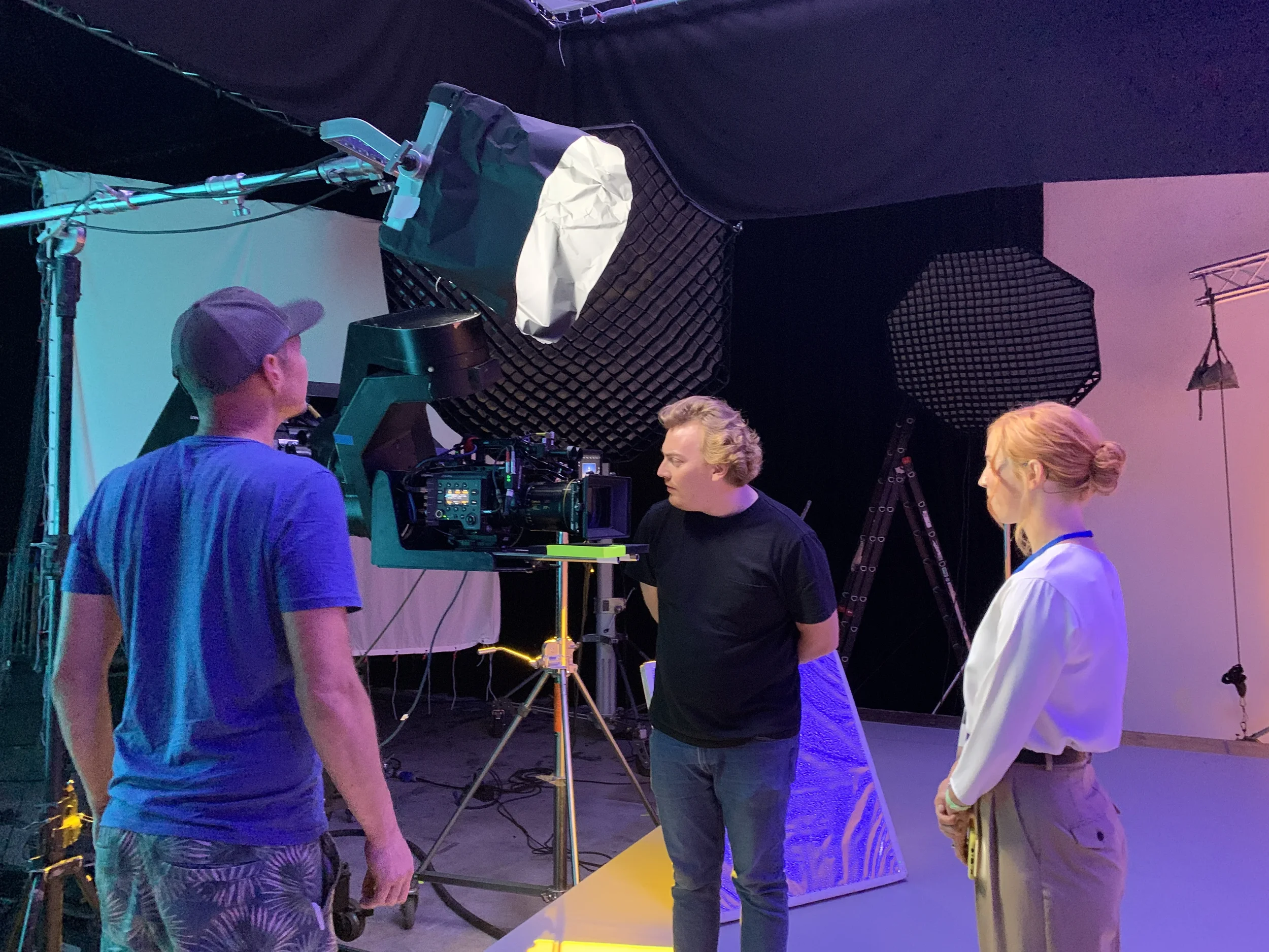 Three people in a studio environment with professional camera equipment, two of them are camera operators and one appears to be a performer or actor engaged in a filming or photography session.