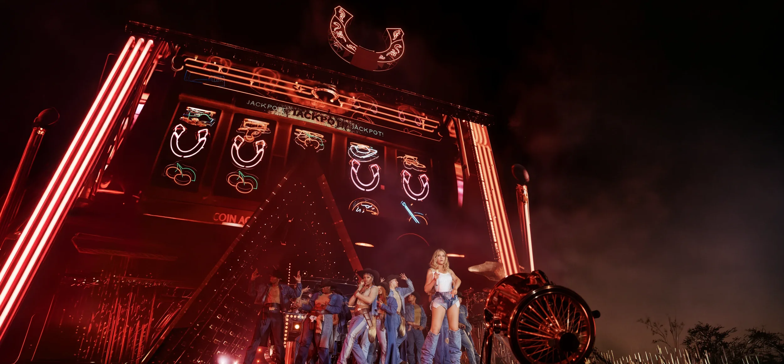 Night scene at a neon-lit amusement ride with performers dressed in cowboy style outfits, performing in front of a large illuminated sign with neon symbols and the word 'JACKPOT'.
