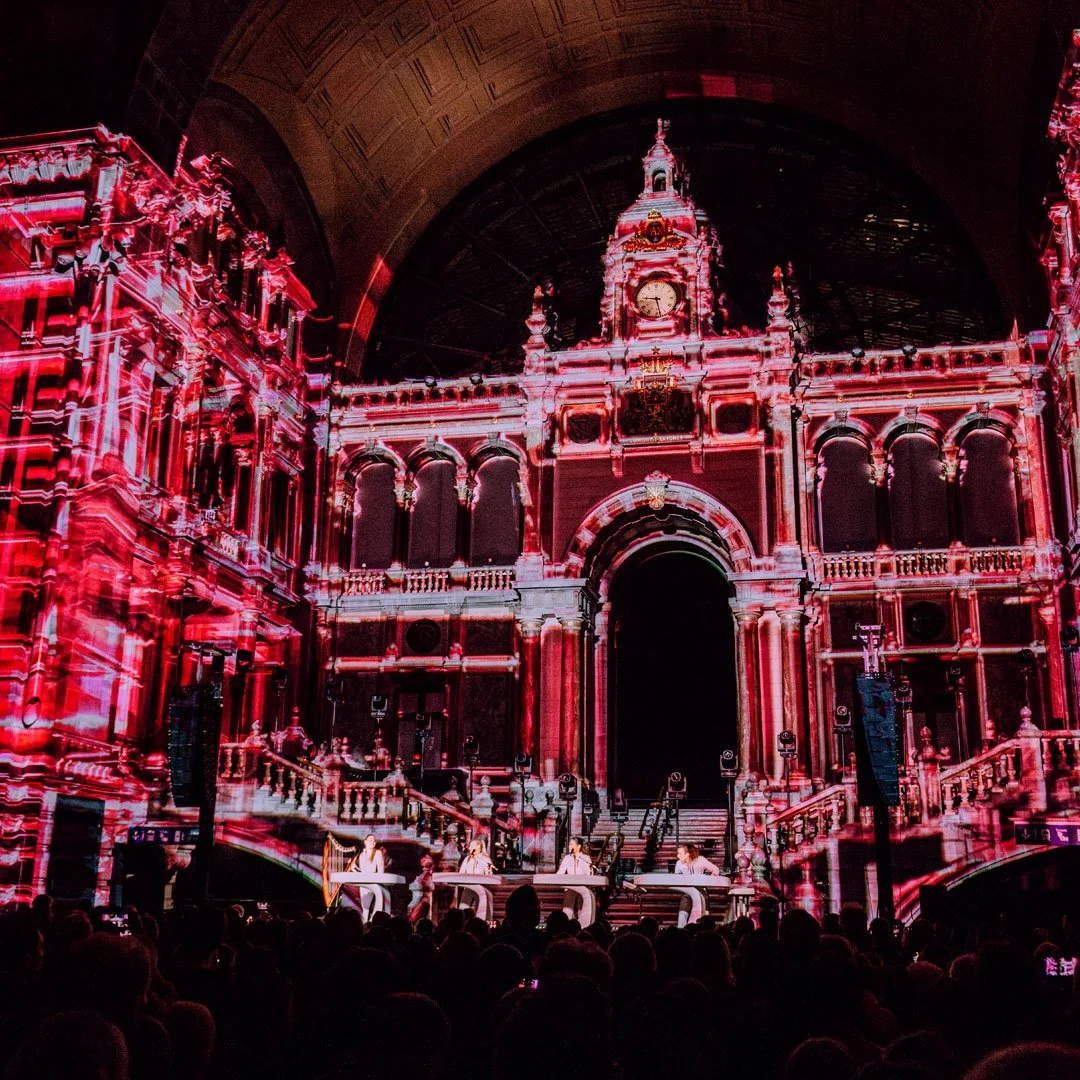 A stage with a projection of a historic building facade, illuminated in pink and red tones, with four performers seated at white tables in front of the building.