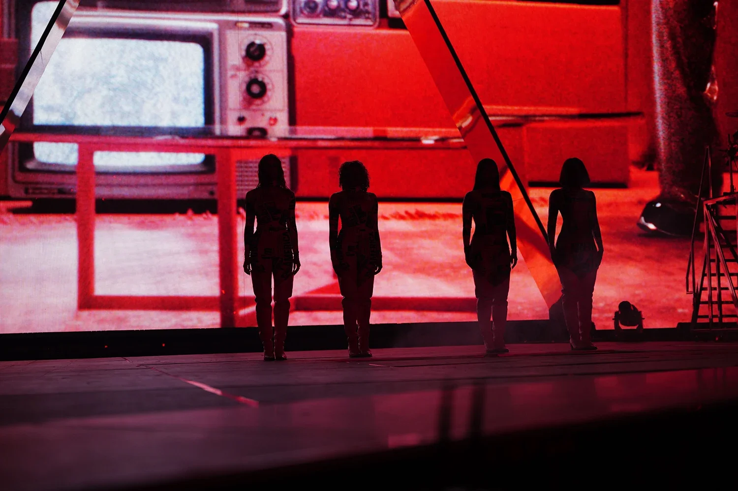 Four silhouetted women standing on stage in front of a large, abstract red and black digital backdrop featuring a vintage television with static screen.