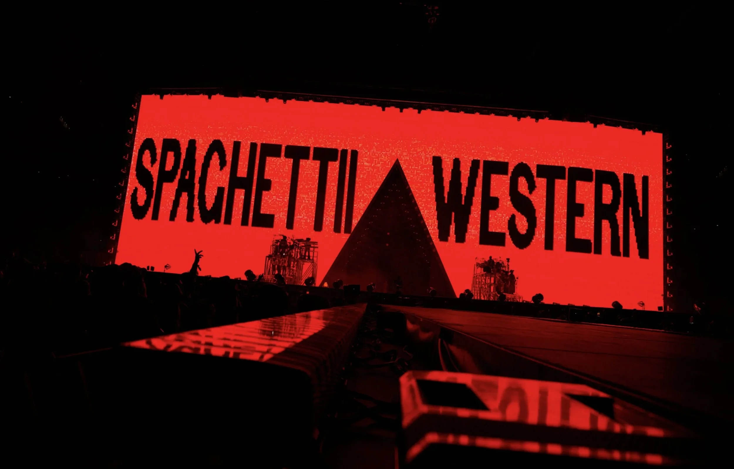 Stage with large red screen displaying the words 'SPACETTI WESTERN' in bold black letters, with a pyramid-shaped structure in the center and silhouettes of people and equipment in the foreground.