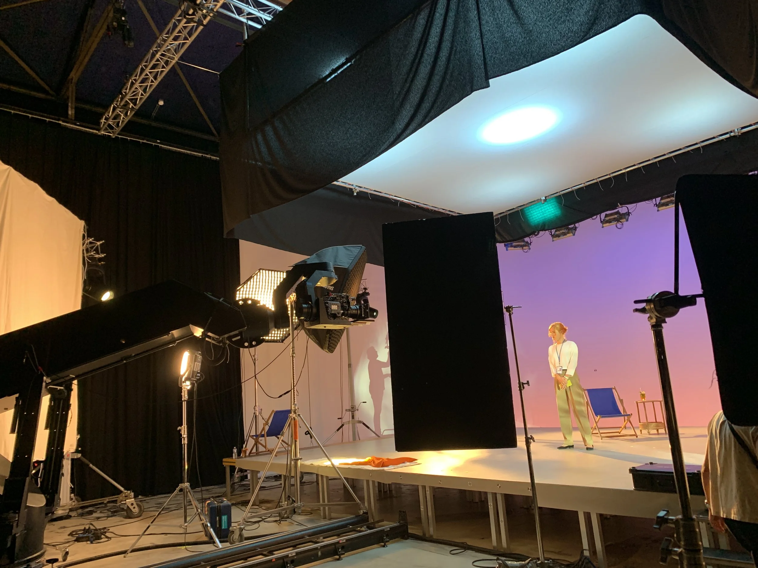 Professional video production set with lighting, cameras, and a stage where a woman stands for filming, with a purple backdrop and beach chair on the stage.
