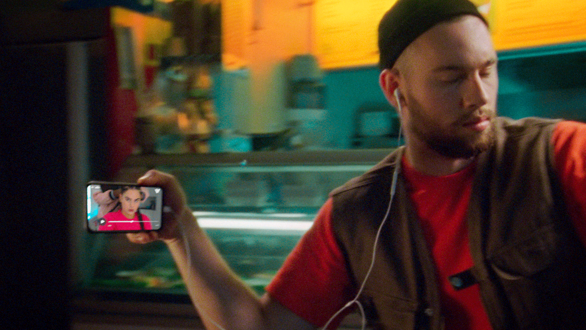A young man with a beard, wearing a black beanie, red shirt, and brown vest, is sitting in a moving vehicle, possibly a bus or train, with his eyes closed and earphones in. He is holding a smartphone in his right hand, capturing his reflection on the