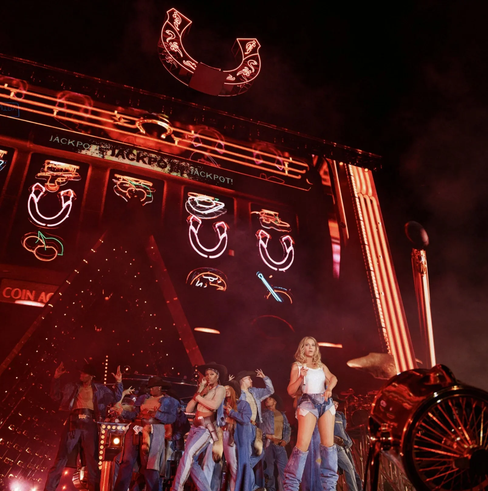 A concert stage with neon lights and signs, featuring a female performer in denim shorts and a white top, surrounded by backup dancers in western-style outfits. The large screen behind displays slot machine symbols, and neon signs like a horseshoe and a revolver are visible, creating a lively, Western-themed atmosphere.
