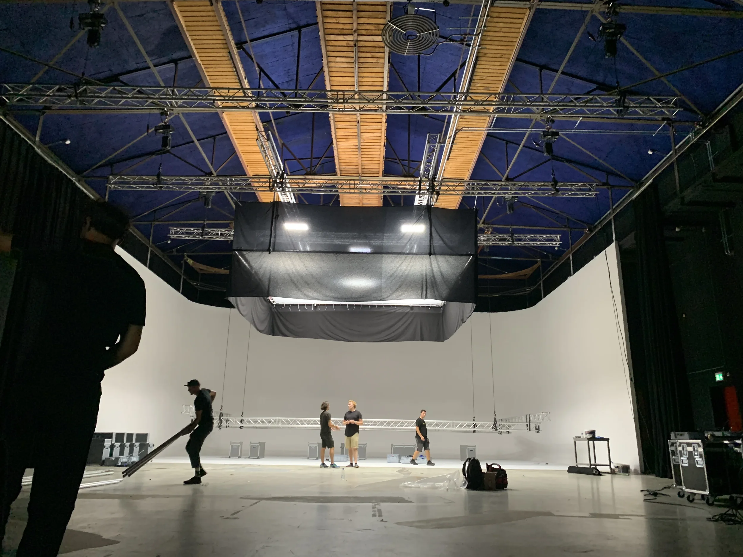 Film or photography studio with a white cyclorama wall, overhead rigging and equipment, and several people setting up or working on the scene.