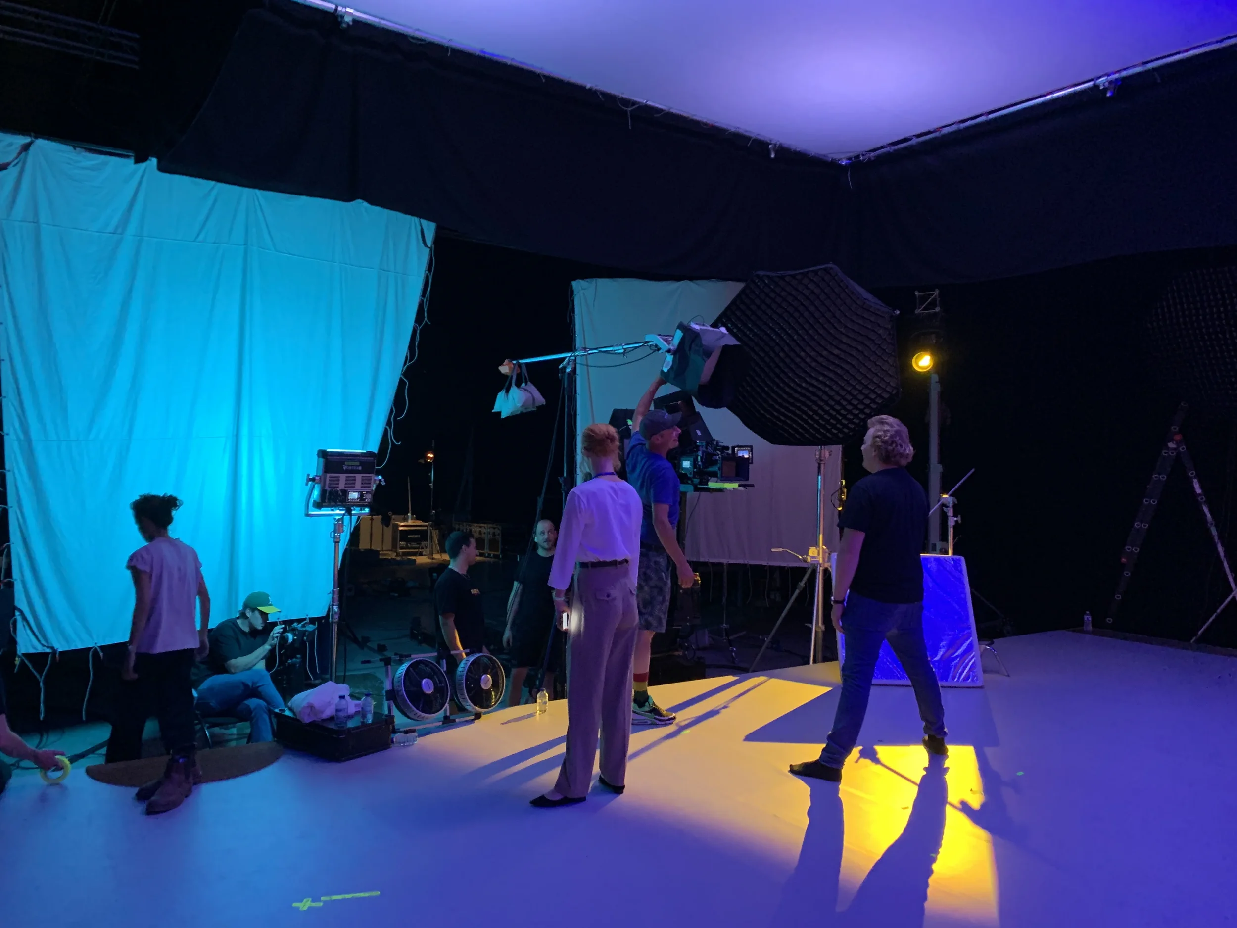 Film crew setting up equipment on a stage with blue and yellow lighting.