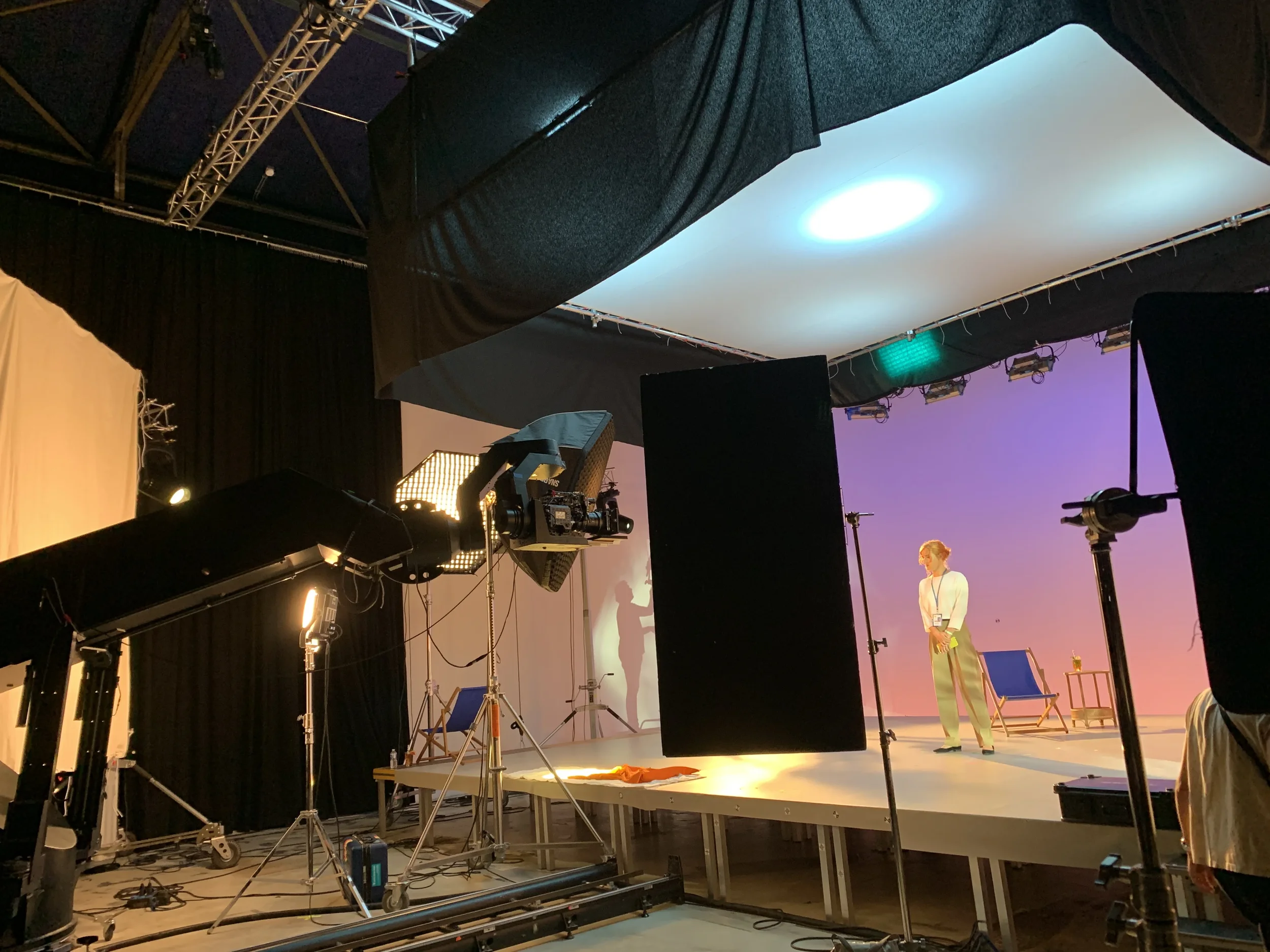 A person stands on a stage with a purple backdrop, surrounded by professional lighting and filming equipment in a studio setting.