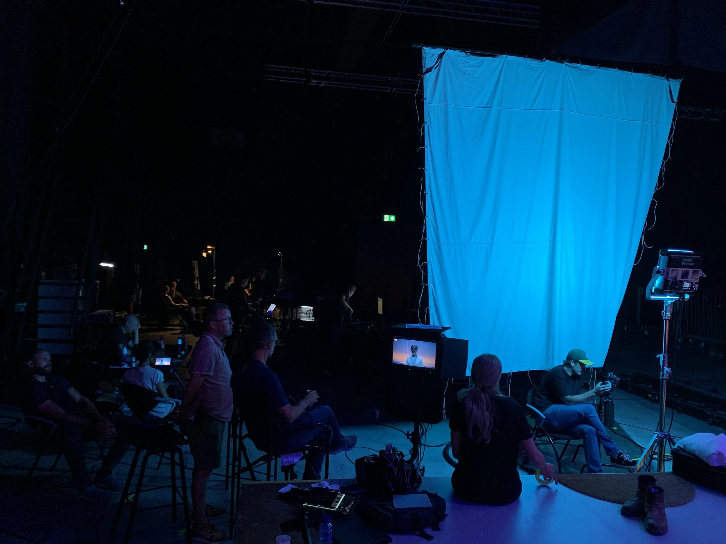 Stage setup with a large white screen, crew members sitting and working in an auditorium, some on chairs, some with laptops, a monitor showing a live feed, and professional lighting equipment.