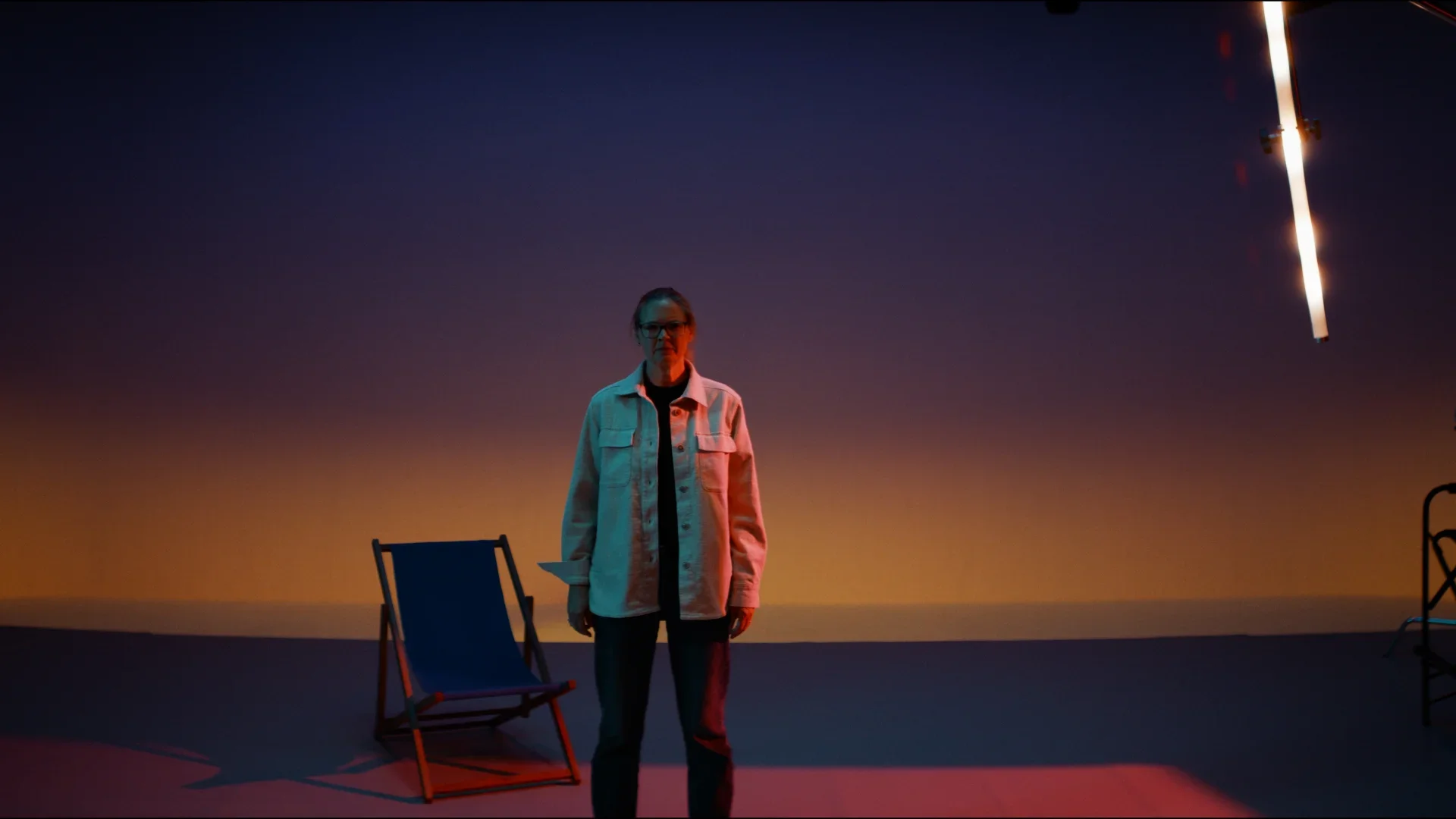 A woman standing on a stage with a colorful gradient background, with chairs around her and a bright streak of light in the top right corner.