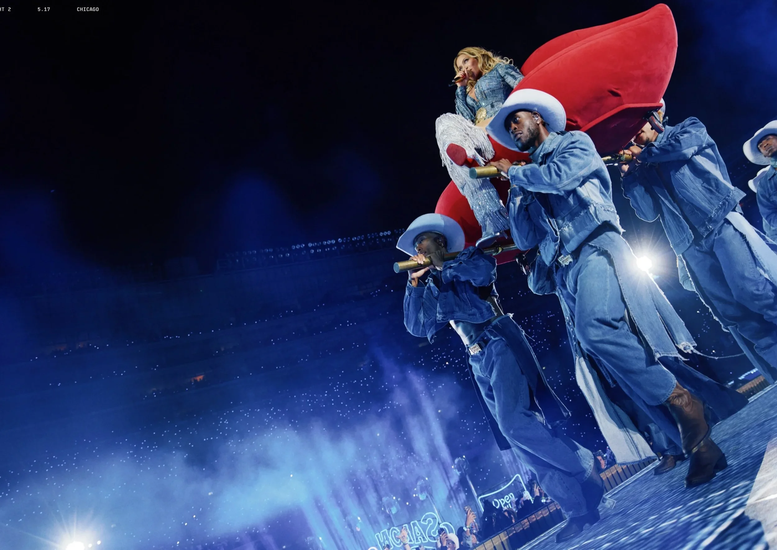 Performer in a sparkling outfit sitting on a large prop, surrounded by dancers in denim outfits with white cowboy hats, performing on a stage at night in Chicago.