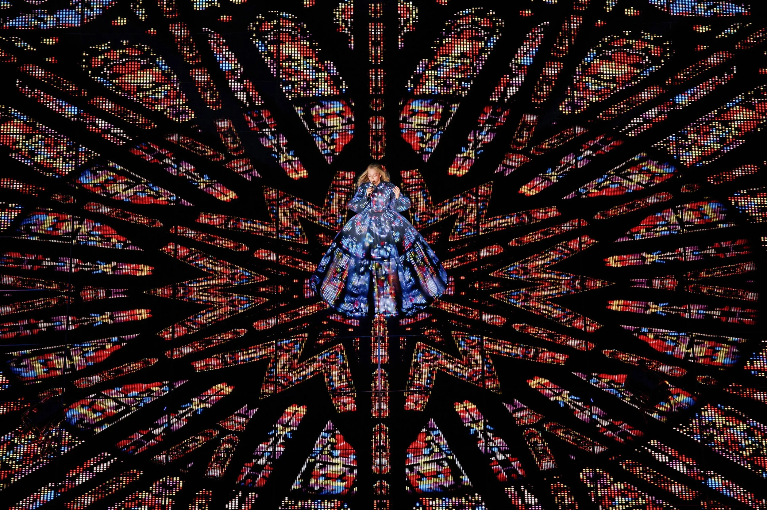 A woman in a long, flowing dress with a colorful geometric pattern stands in the center of a vibrant, illuminated mosaic background resembling stained glass.