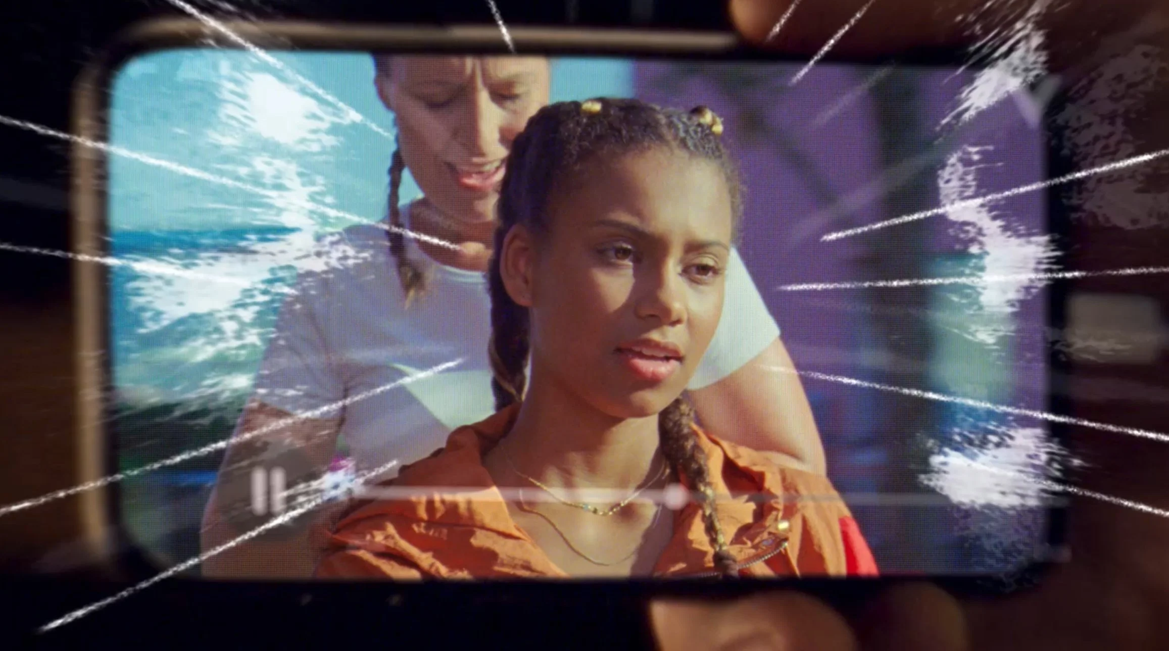 A woman with braided hair and an orange jacket on a video call with a young girl with braided hair, wearing an orange hoodie. The screen appears to be cracked.