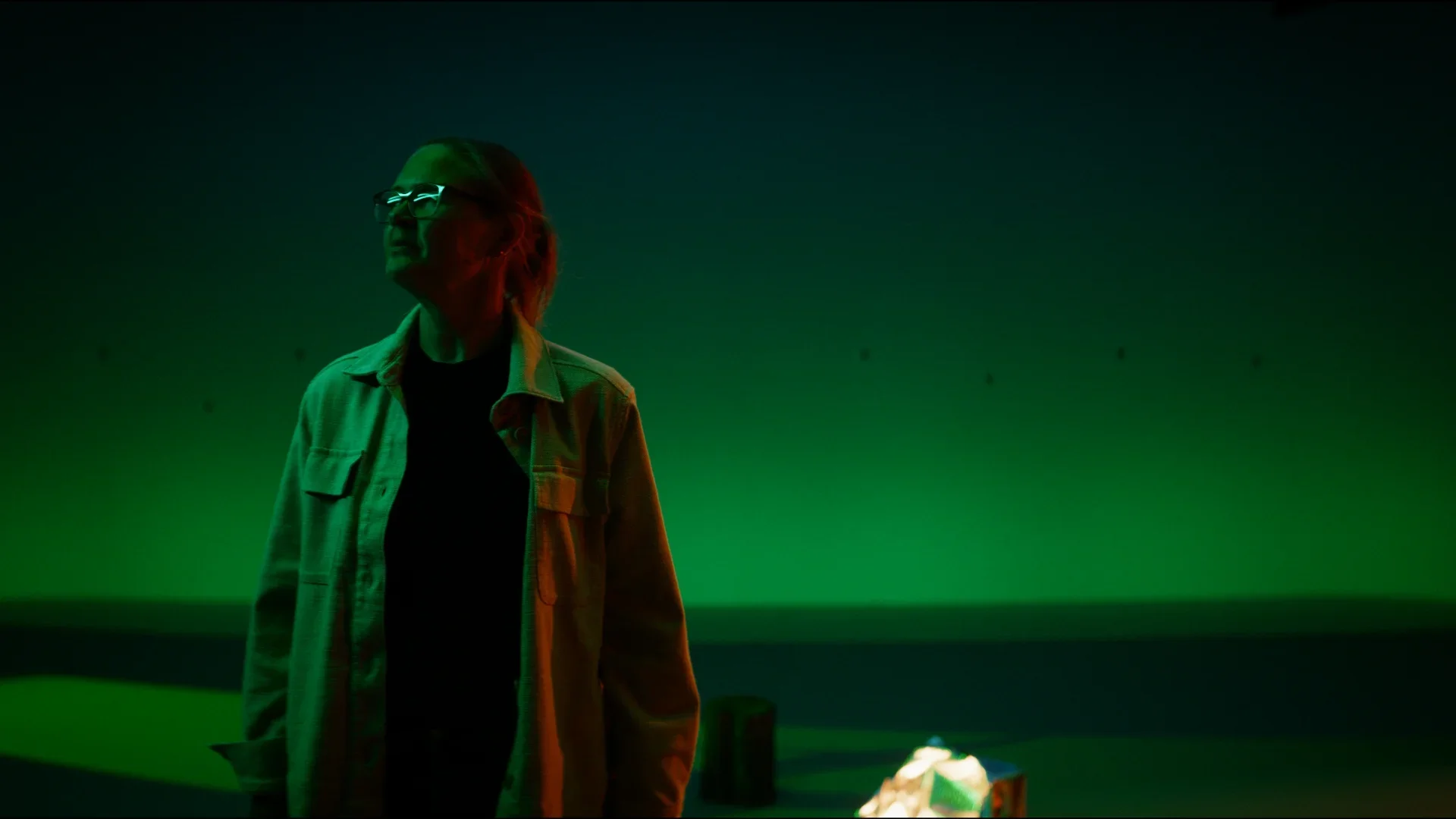 A woman with glasses and short hair stands in a dark, green-illuminated space with a faint horizon line. She is wearing a light-colored jacket over a dark shirt and looks to the left.