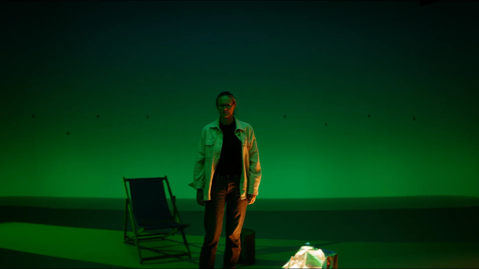 A woman stands in a dimly lit room with green lighting, a chair, and a bag on the floor, with walls that have small decorations or holes.