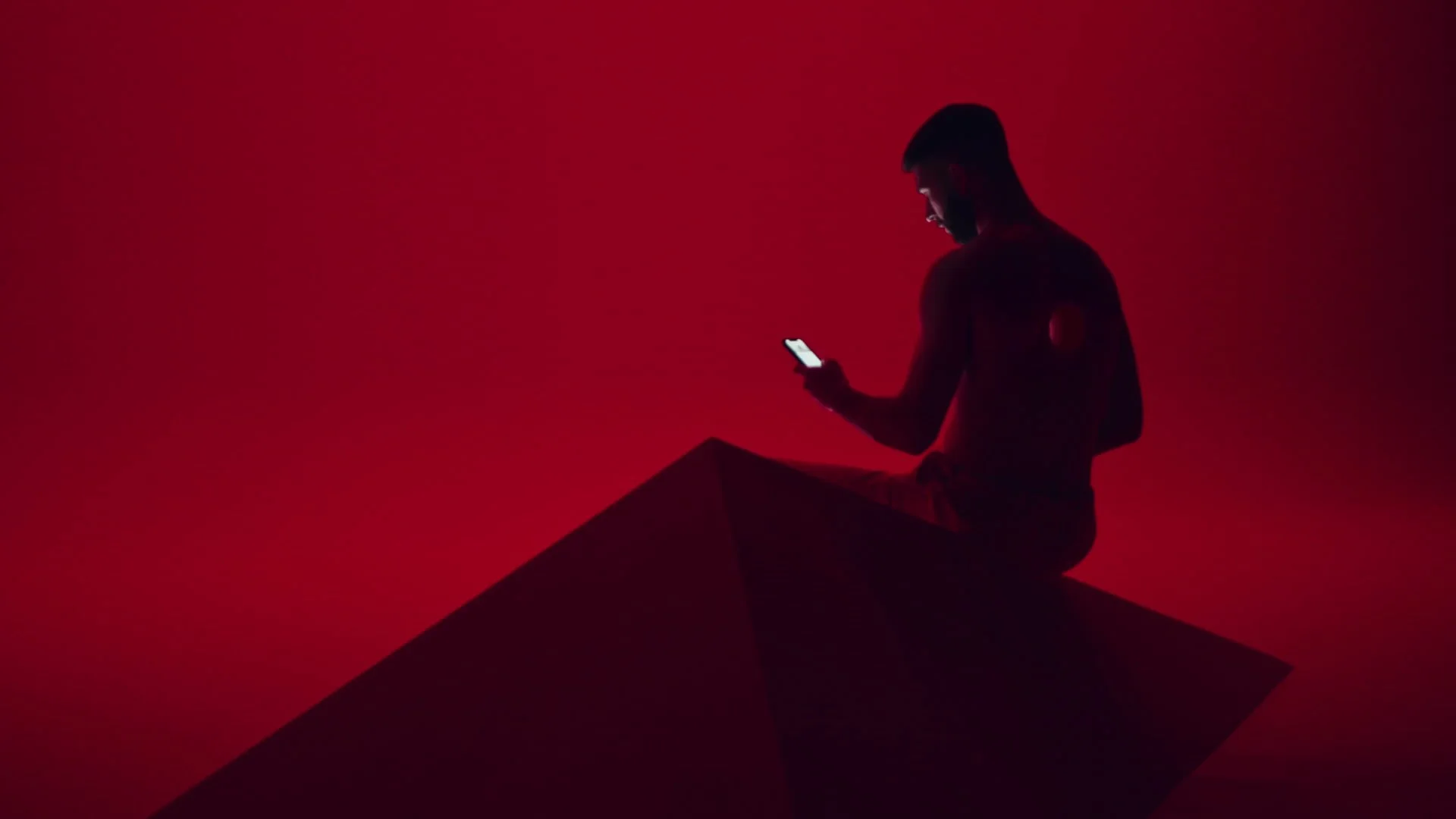 Silhouette of a man sitting on a dark geometric shape, looking at his phone against a red background.