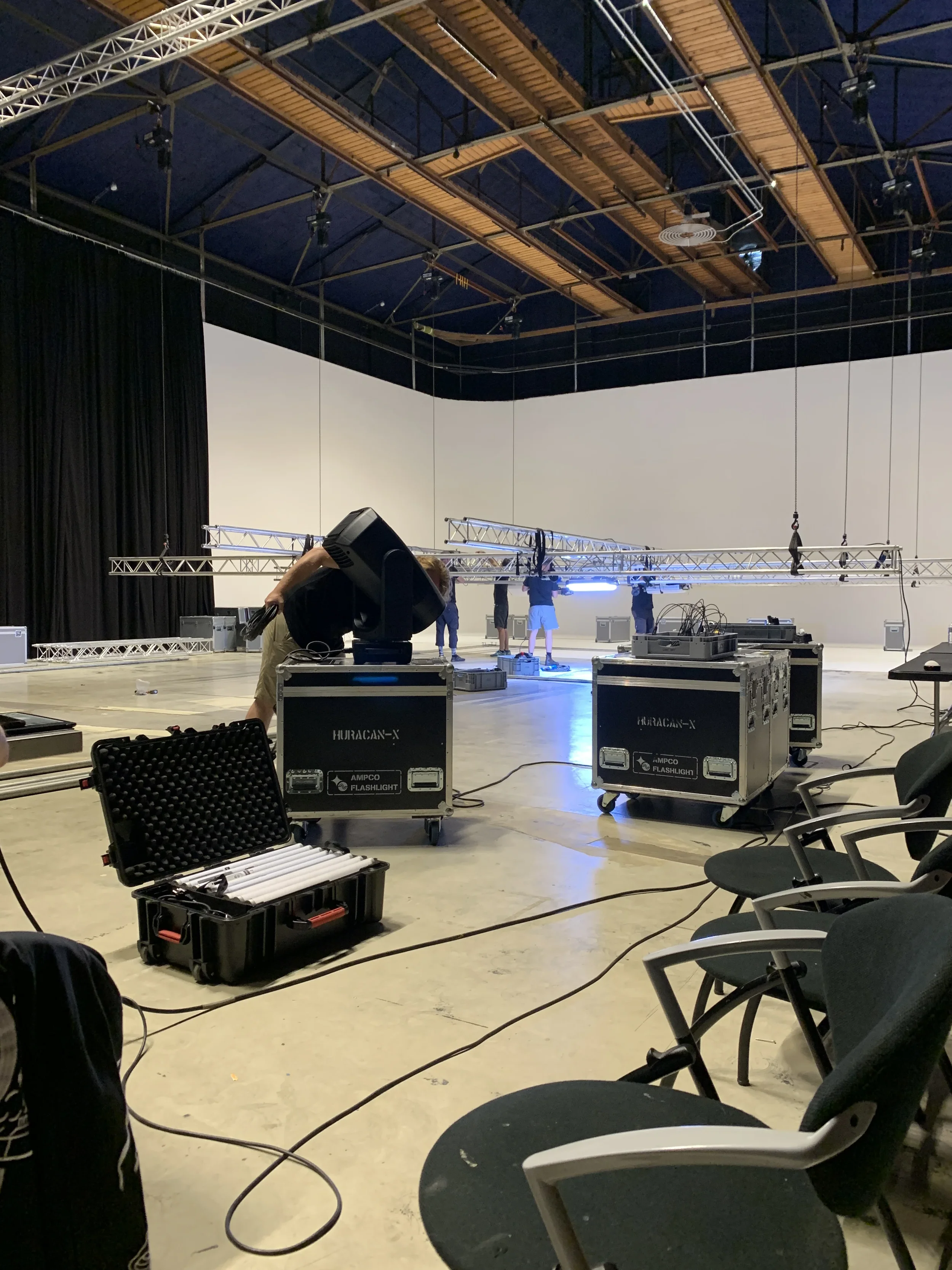 A large, empty indoor stage setup with lighting and support structures, crew members working, and chairs arranged on the side.