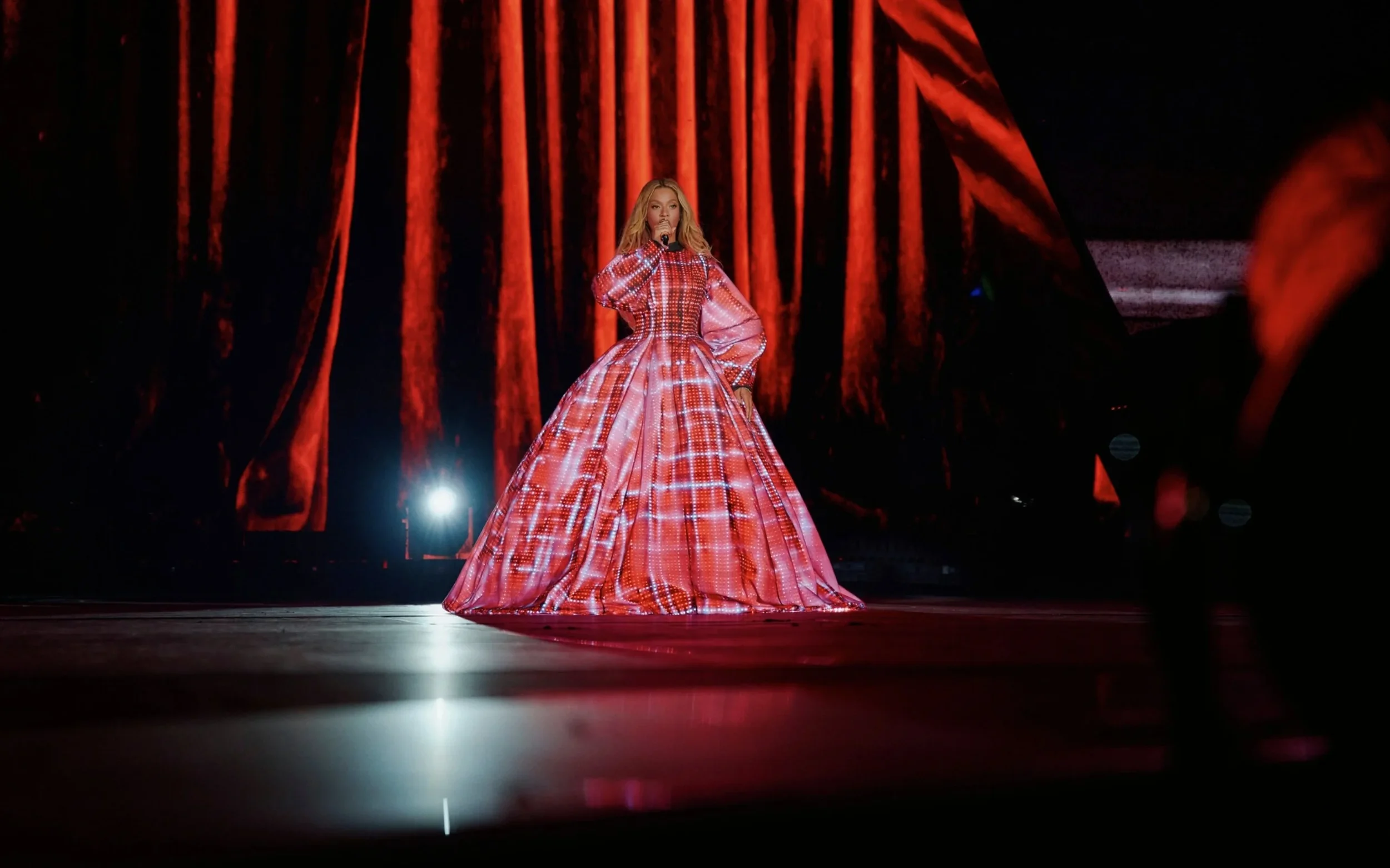 Taylor Swift performing on stage in a red plaid dress with a full skirt and puffy sleeves, standing before a large red curtain, illuminated by stage lights.
