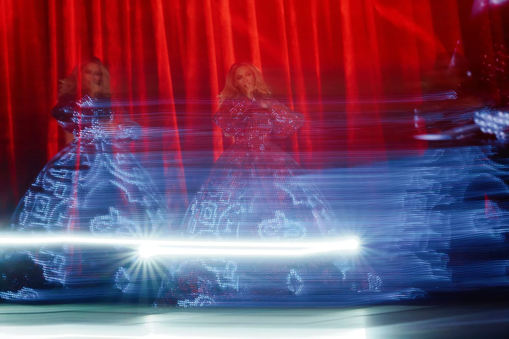 Performance of a singer on stage with red curtains in the background, wearing a shiny, embellished dress, with light streaks creating a dynamic visual effect.