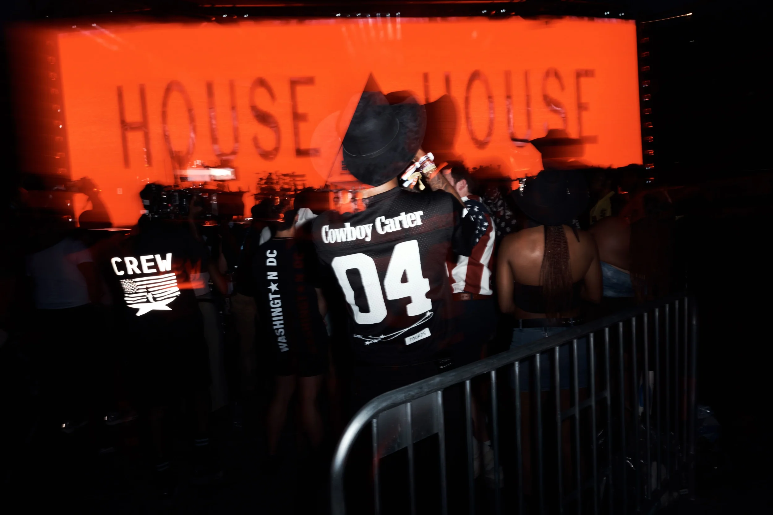 People attending a House of House concert or event, with a large orange screen displaying 'HOUSE OF HOUSE' in the background.