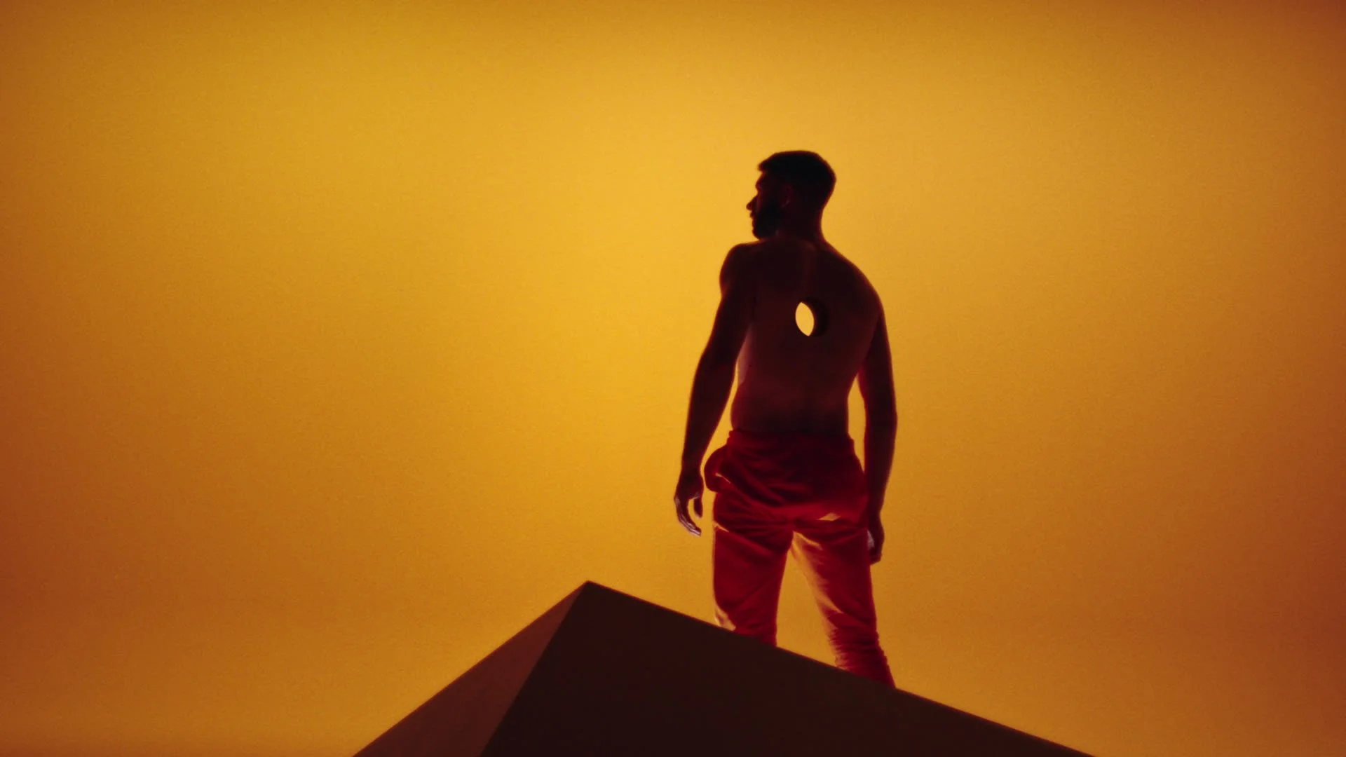 Silhouette of a shirtless man with a hole in his chest, standing on a pyramid-shaped structure against an orange background.