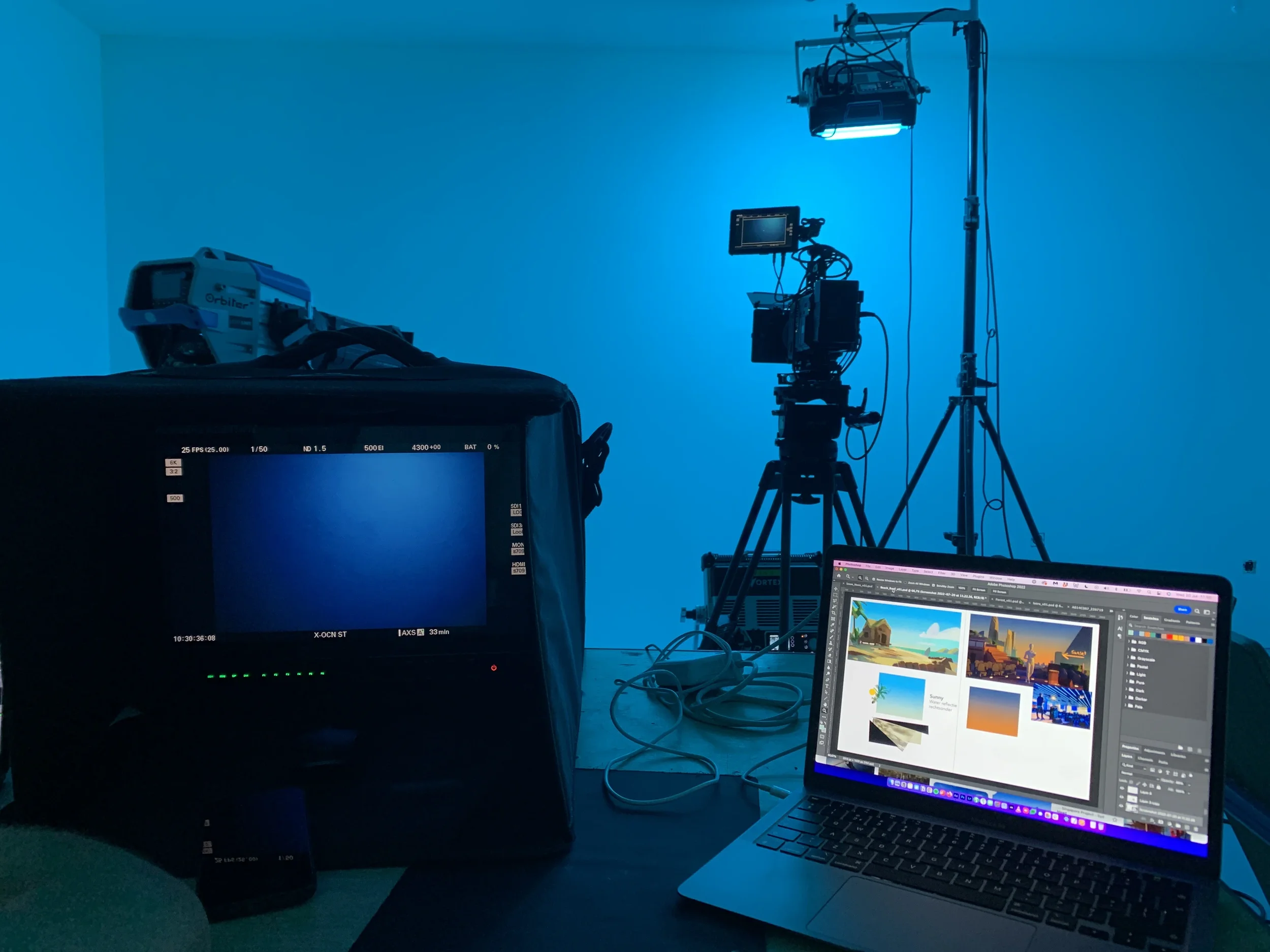 A professional video or photo shoot setup with a camera on a tripod, an LED light, a monitor, and a laptop open to a design application, all in a blue-lit studio environment.