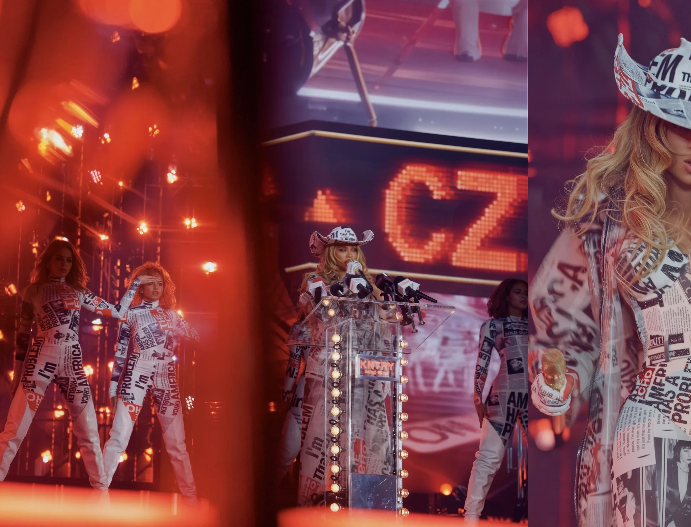 A woman dressed in newspaper print outfit, wearing a matching hat, is speaking at a transparent podium covered with microphone devices during a stage event. Behind her are three women in similar newspaper print outfits, one of whom has her hand on th