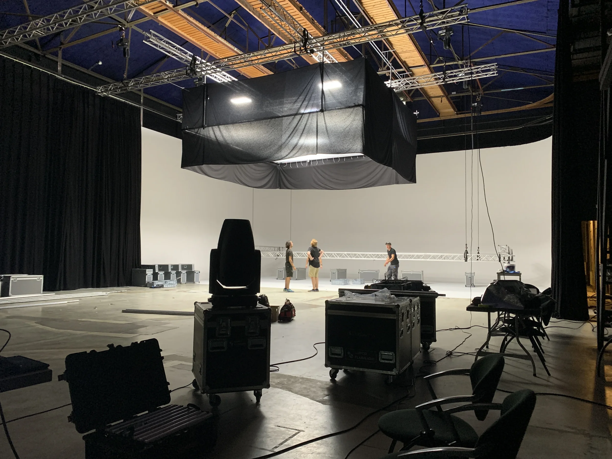 Empty film or photo studio set with three people working, large white backdrop, overhead lighting and rigging, black curtains on sides, various equipment and cables in the foreground.