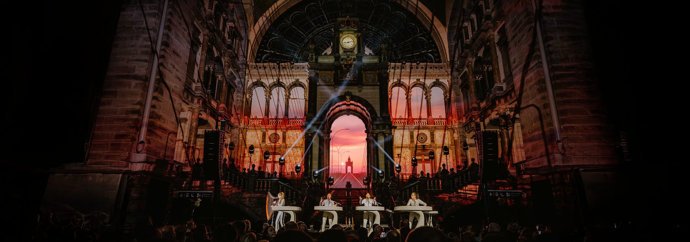 Concert inside a historic building with elaborate architecture, sunset sky seen through large arched windows, four performers at white desks, stage lighting, and an audience in the foreground.