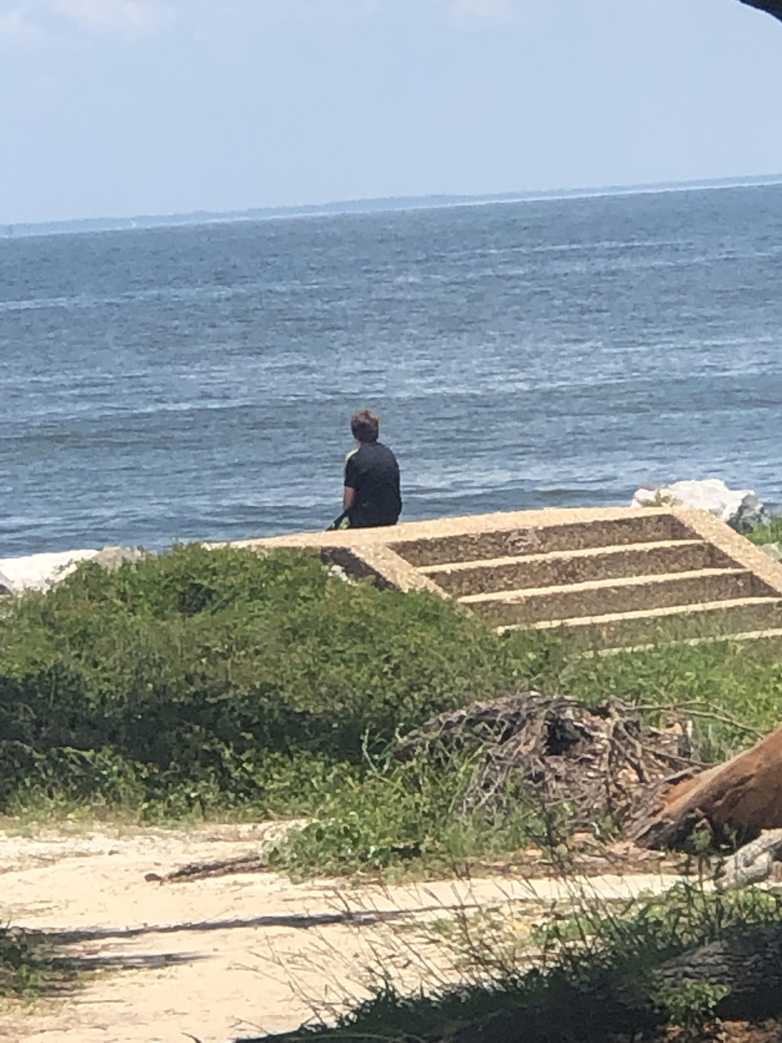 There's a young man sitting on a concrete wall in the center of the picture. In the foreground is sand with greenery and a brush pile and in the background the wide ocean. The young man appears to be deep in thought.