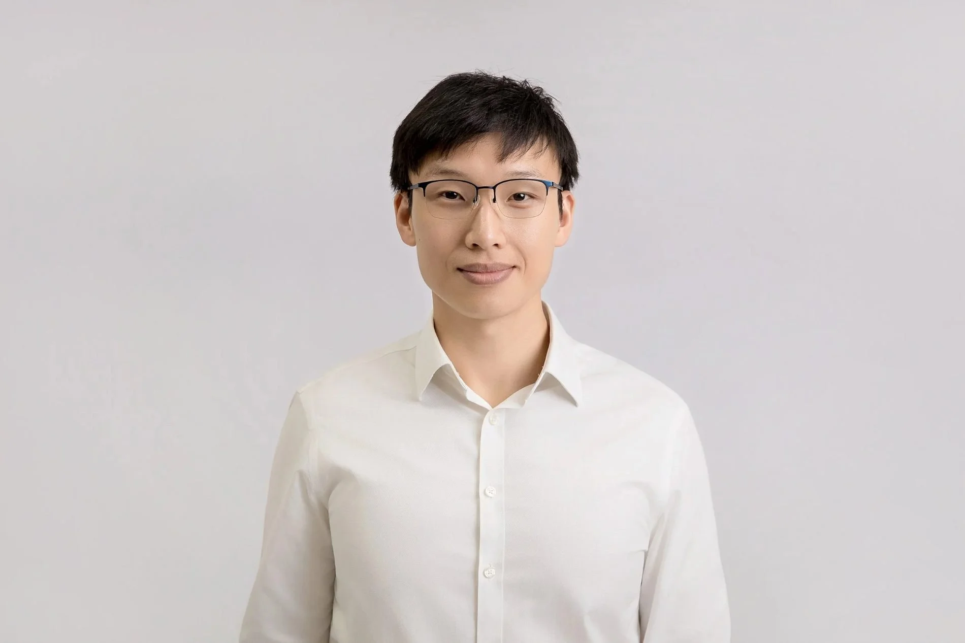 A portrait of a young Asian man with short black hair, glasses, and a slight smile, wearing a white dress shirt against a plain light gray background.
