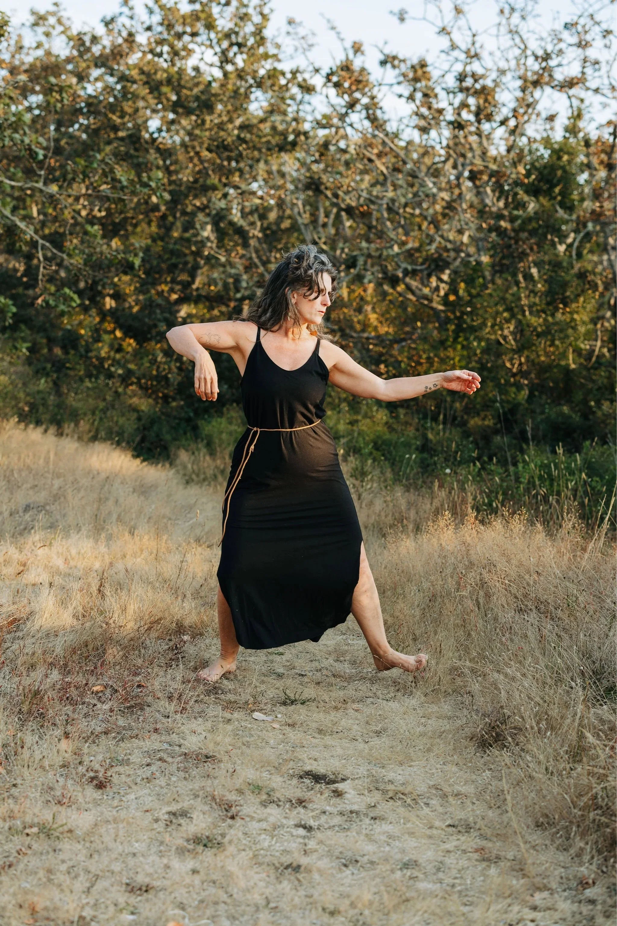 A woman in a black dress dances outdoors in a field with tall grass and a backdrop of trees.
