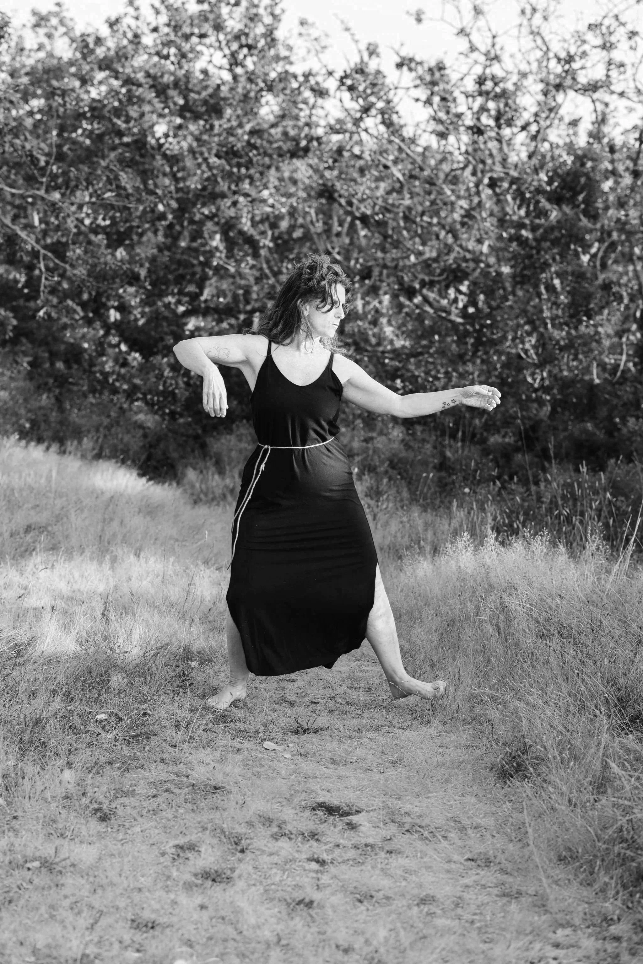 A woman in a black dress dances outdoors in a field with tall grass and a backdrop of trees.