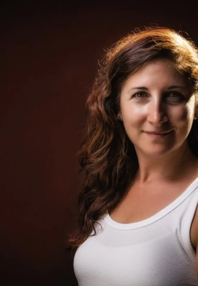 Woman with long wavy hair wearing a white tank top, smiling at the camera, with a dark background.