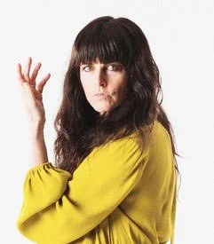 A woman with long dark hair wearing a yellow top poses with her hand raised against a plain white background.