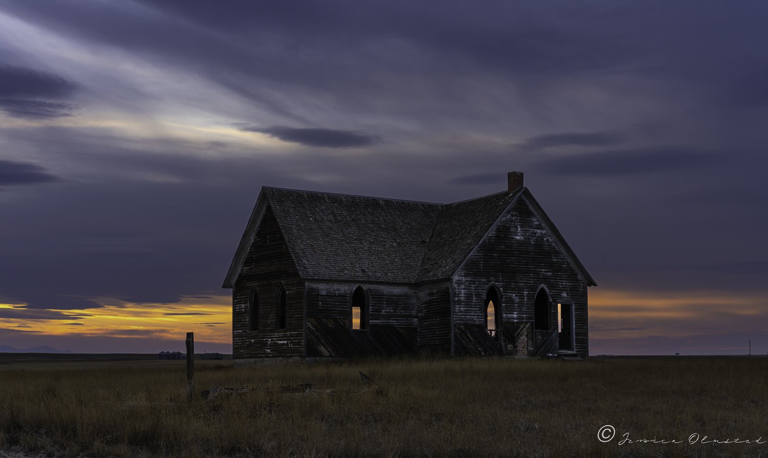 Abandoned wooden church with gothic windows at sunset in a grassy field.