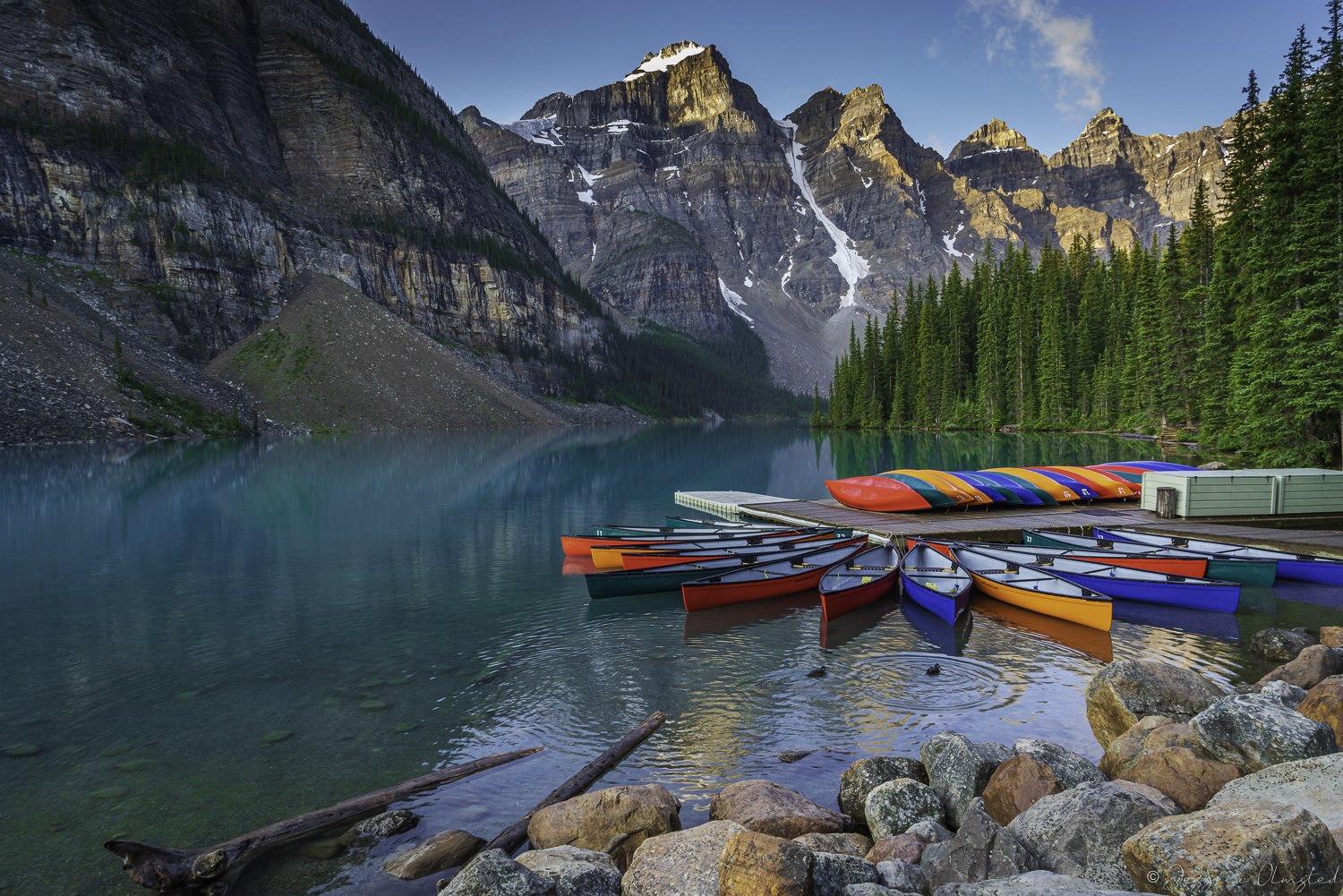 Colorful canoes on a dock at Moraine Lake with rocky mountains and pine trees in the background.