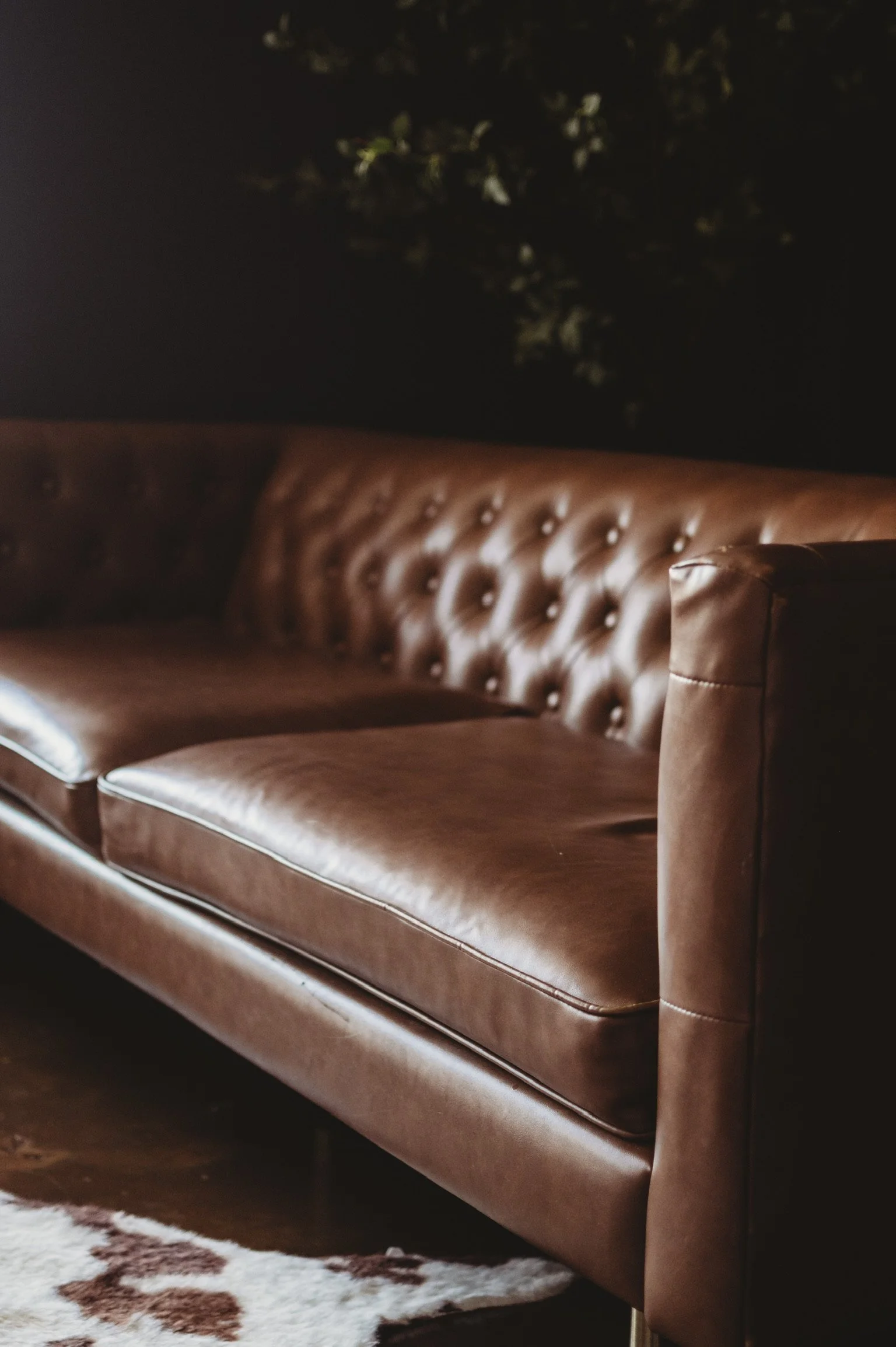 A brown leather sofa with button-tufted backrest in a dimly lit room. Part of a white and brown cowhide rug is visible in the foreground, and a dark background with some tree branches is faintly seen.