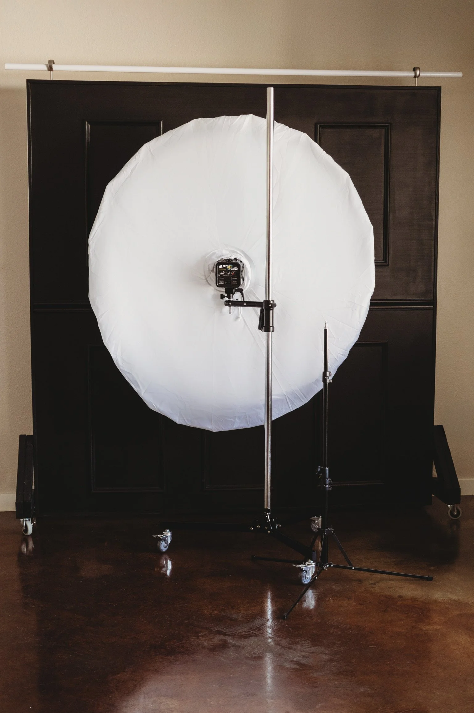 Photography lighting setup with a large white umbrella umbrella and a studio light on a black background with wheels on a hardwood floor.