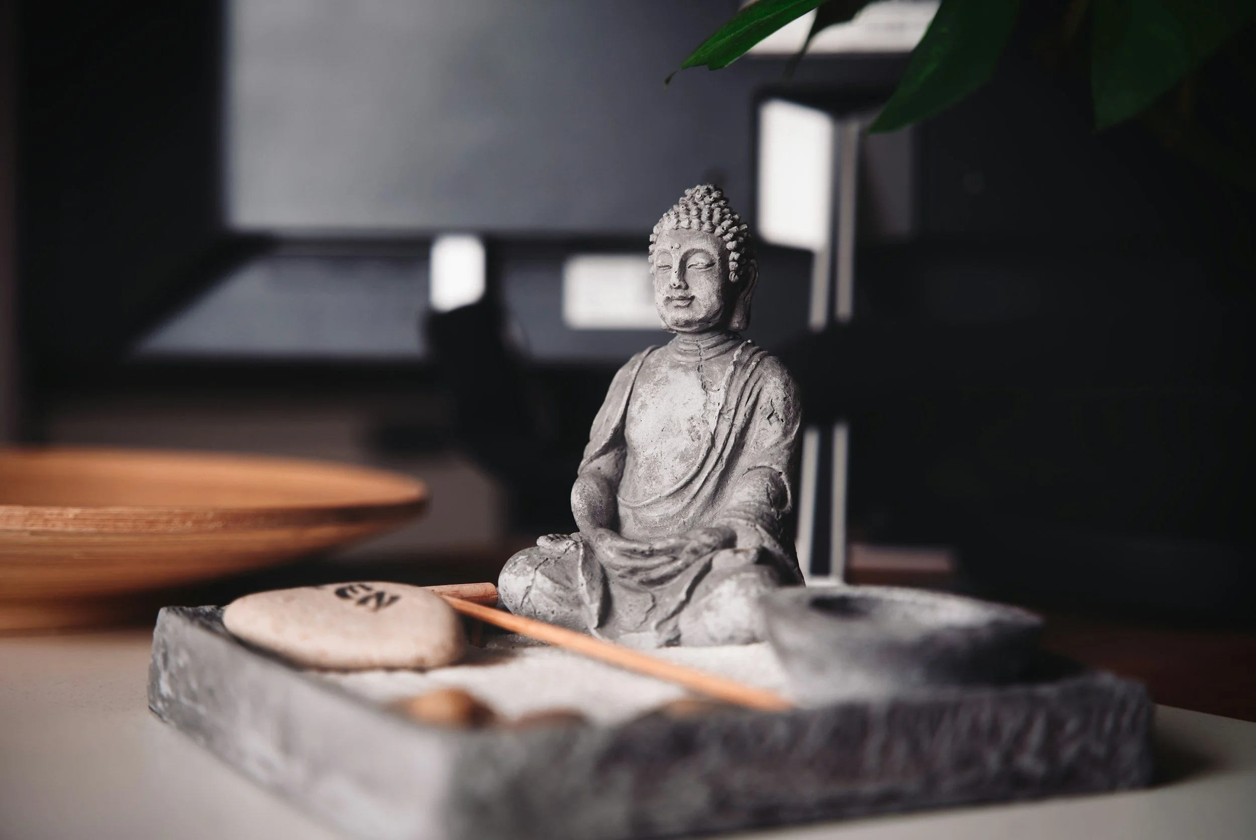 Small Buddha statue in a zen garden setting with rocks, sand, and a wooden paddle on a tray.