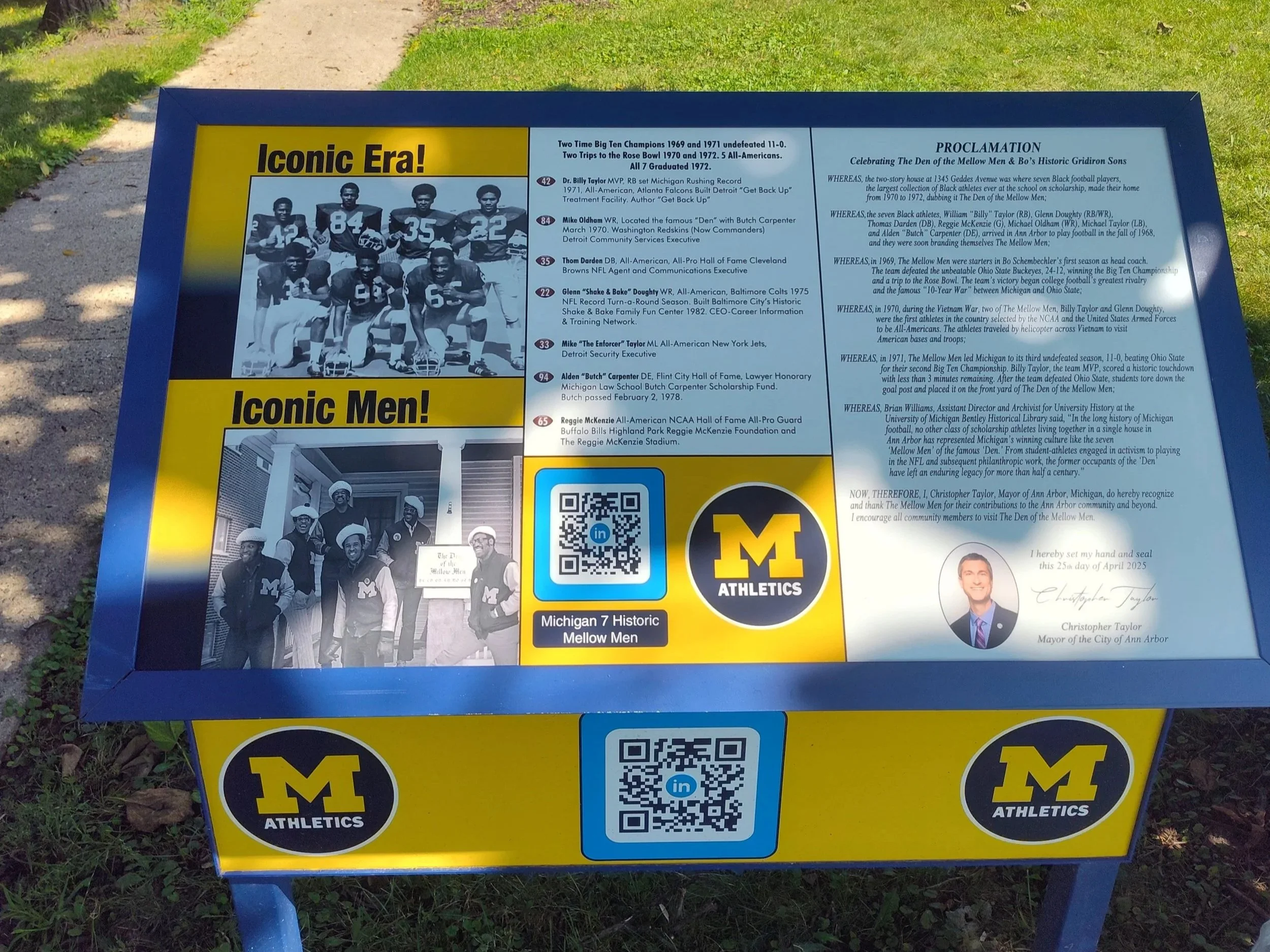 Informational custom signboard celebrating Michigan Wolverines football players and coaches from the 1960s and 1970s, with historical photos and a plaque recognizing their achievements, featuring the U of M Athletics logo.