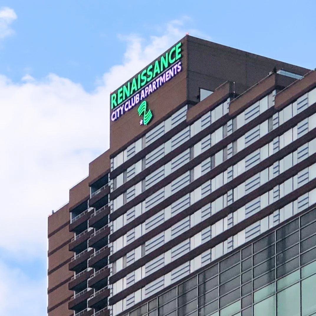 High-rise building with illuminated sign reading 'Renaissance City Club Apartments' and a logo, with a cloudy sky in the background.