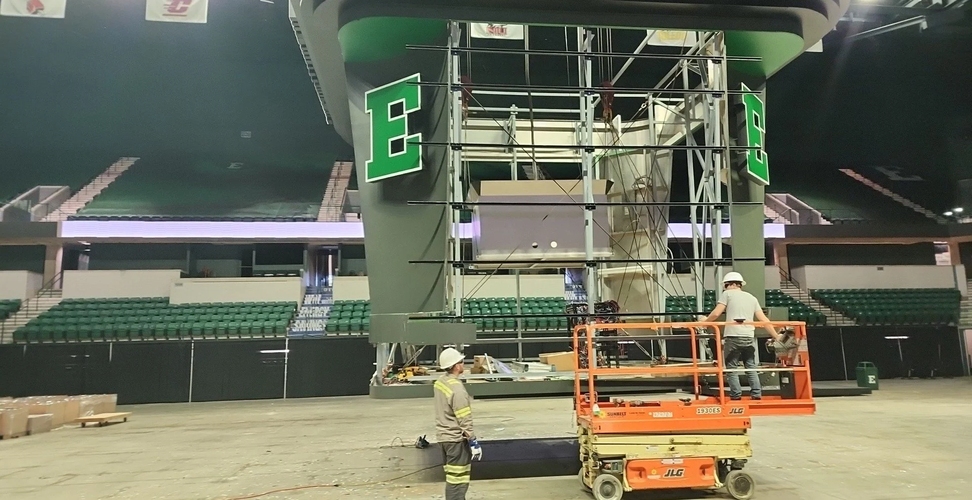 Workers installing a large Eastern Michigan University video scoreboard with white letter E on a green background inside an empty arena.