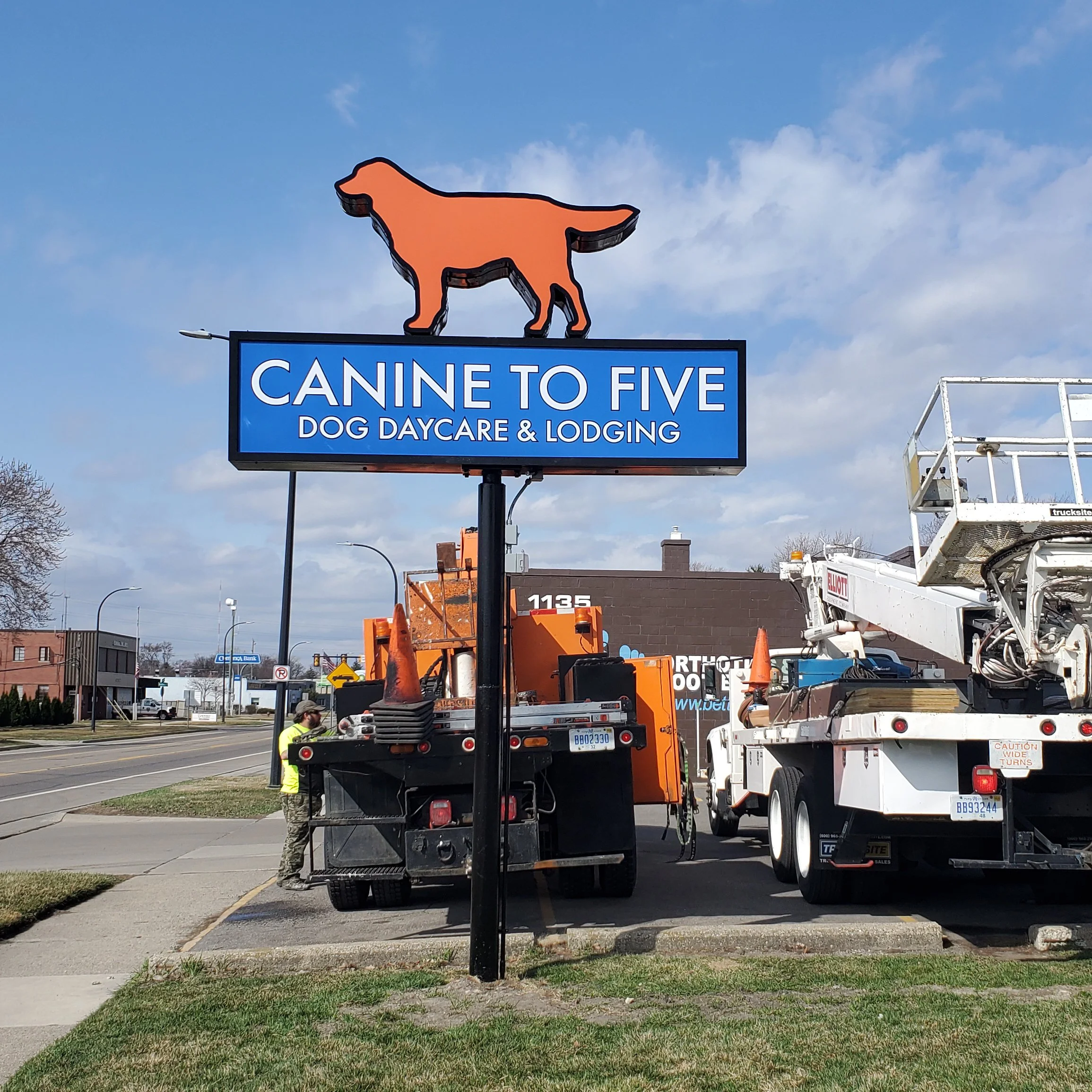 Custom sign for 'Canine to Five' Dog Daycare with a silhouette of a dog on top, situated in a parking lot near street and construction vehicles.