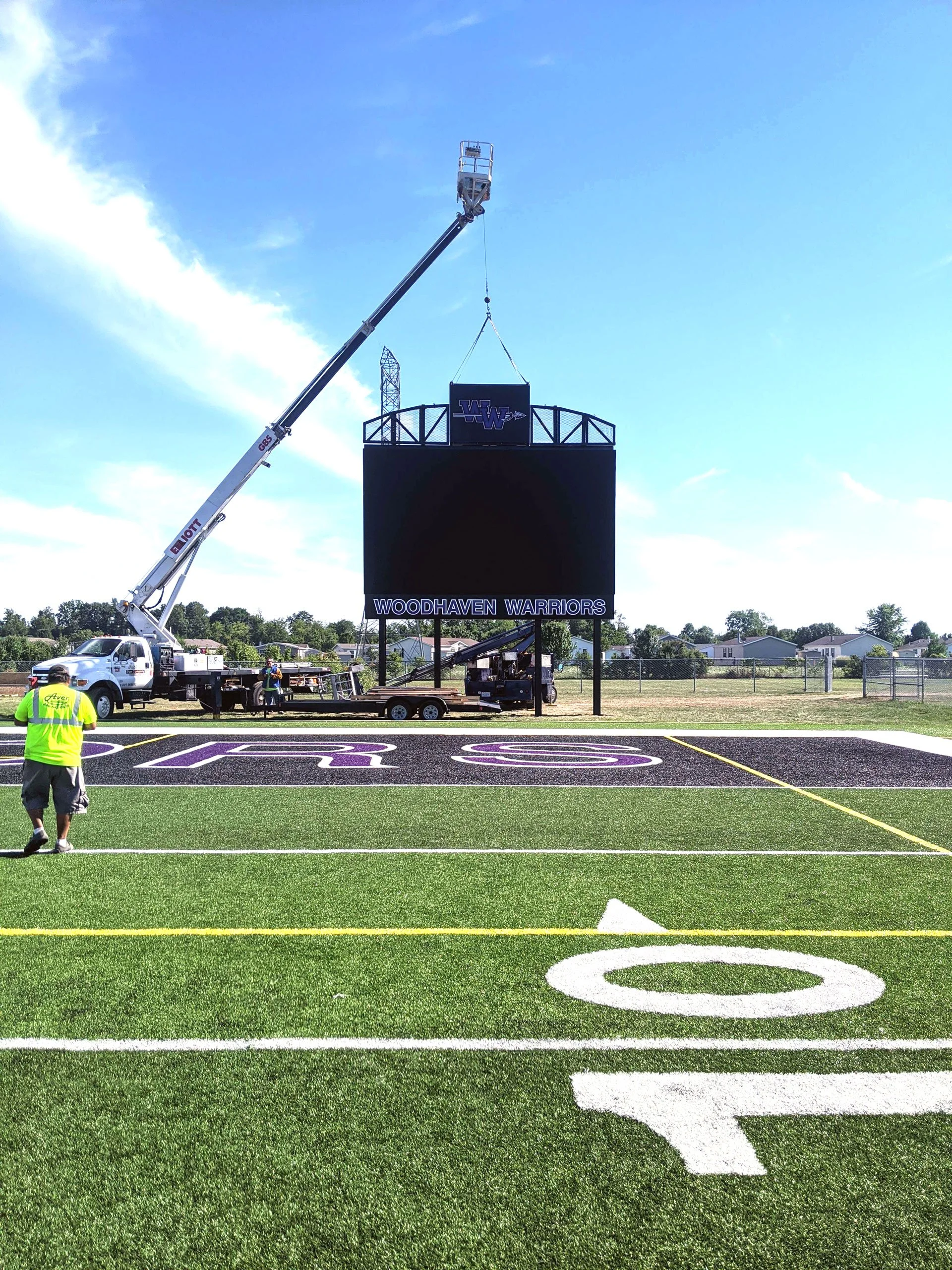 A large custom signage electronic scoreboard on a football field with the words 'WOODHAVEN WARRIORS' at the bottom. A crane is lifting the scoreboard into place. 