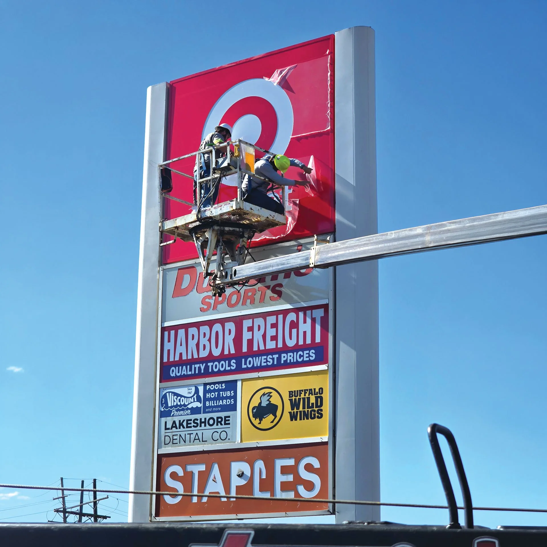 Two workers are on a cherry picker repairing or installing a sign for Target. The sign is tall, with a red and white logo at the top, and the sky is clear and blue.