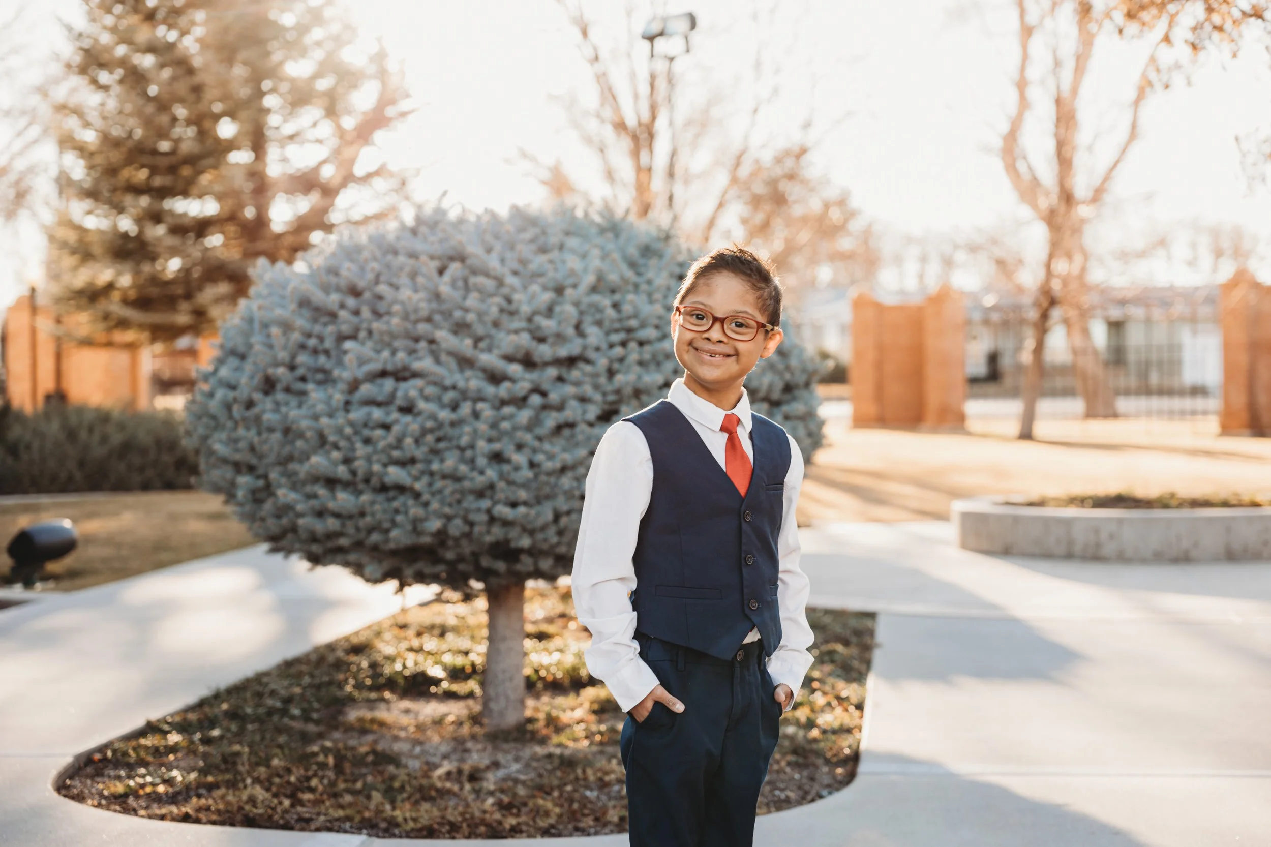 A young boy in a school uniform with glasses, white shirt, navy vest, and red tie, standing outdoors in front of a rounded bush and trees during late afternoon or early evening, smiling at the camera.
