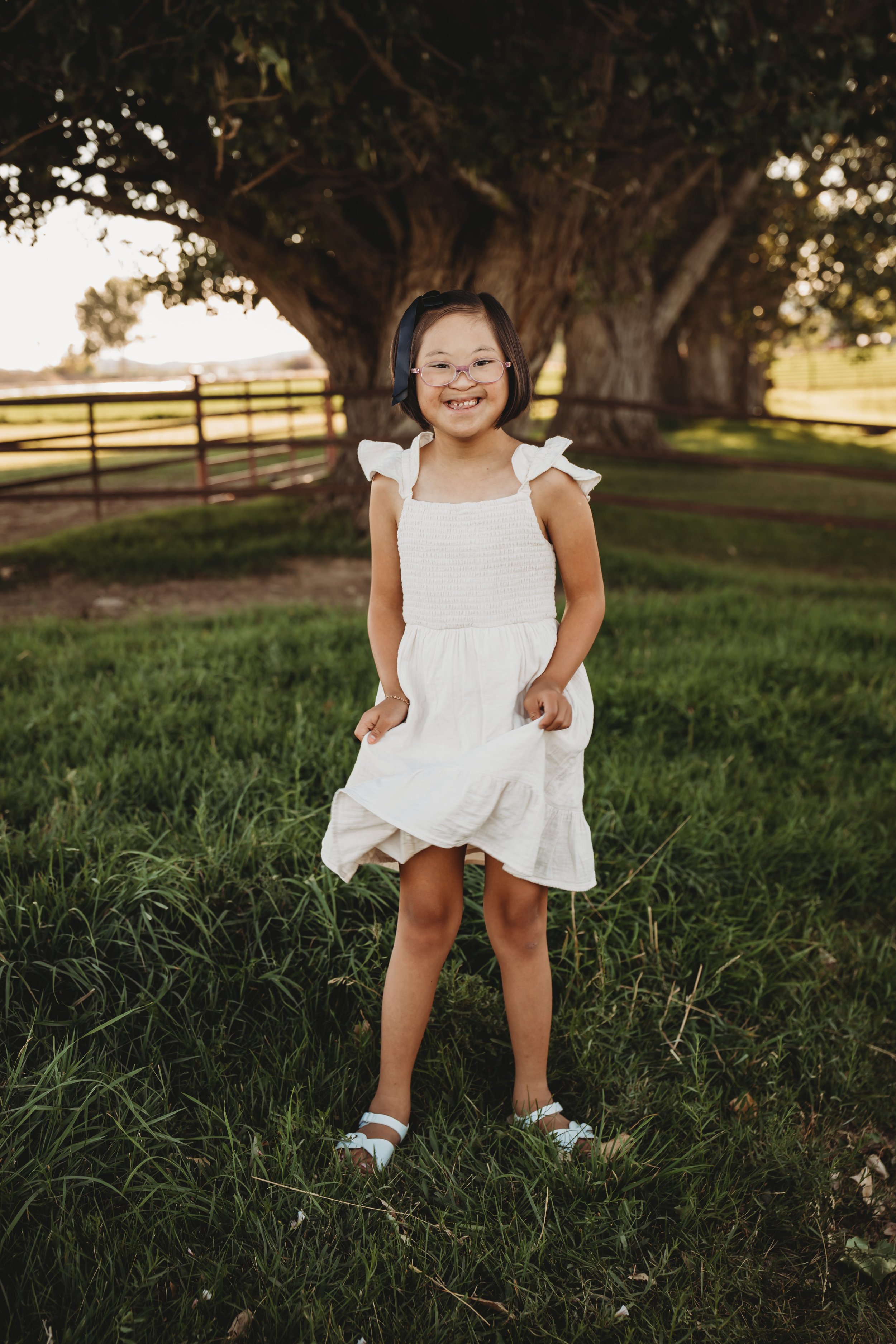 Young girl standing outdoors on grass with a large tree and fencing in the background, smiling at the camera.