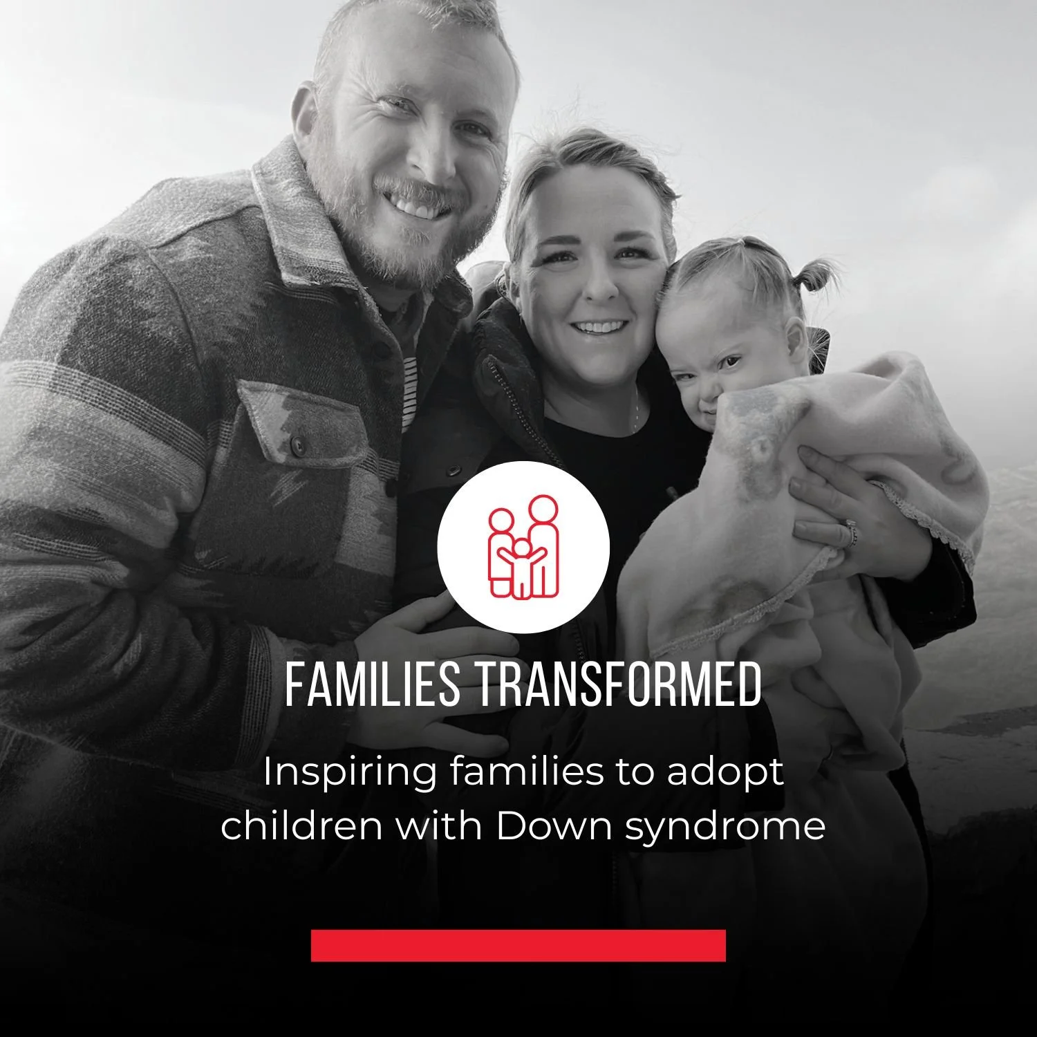 A black and white photo of a happy family of three, including a man, a woman, and a young girl, outdoors. The woman is holding the girl in her arms. Overlaid on the image is a logo with two stylized figures and the text 'FAMILIES TRANSFORMED' and a tagline 'Inspiring families to adopt children with Down syndrome'.