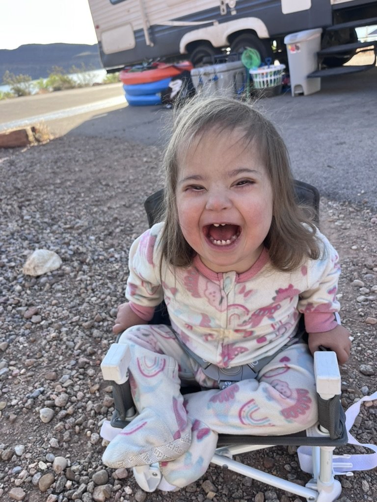 A young girl with down syndrome smiling and laughing while sitting outdoors in a wheelchair on a gravel surface, wearing pajamas with rainbows and unicorns, with a camper trailer and outdoor supplies in the background.